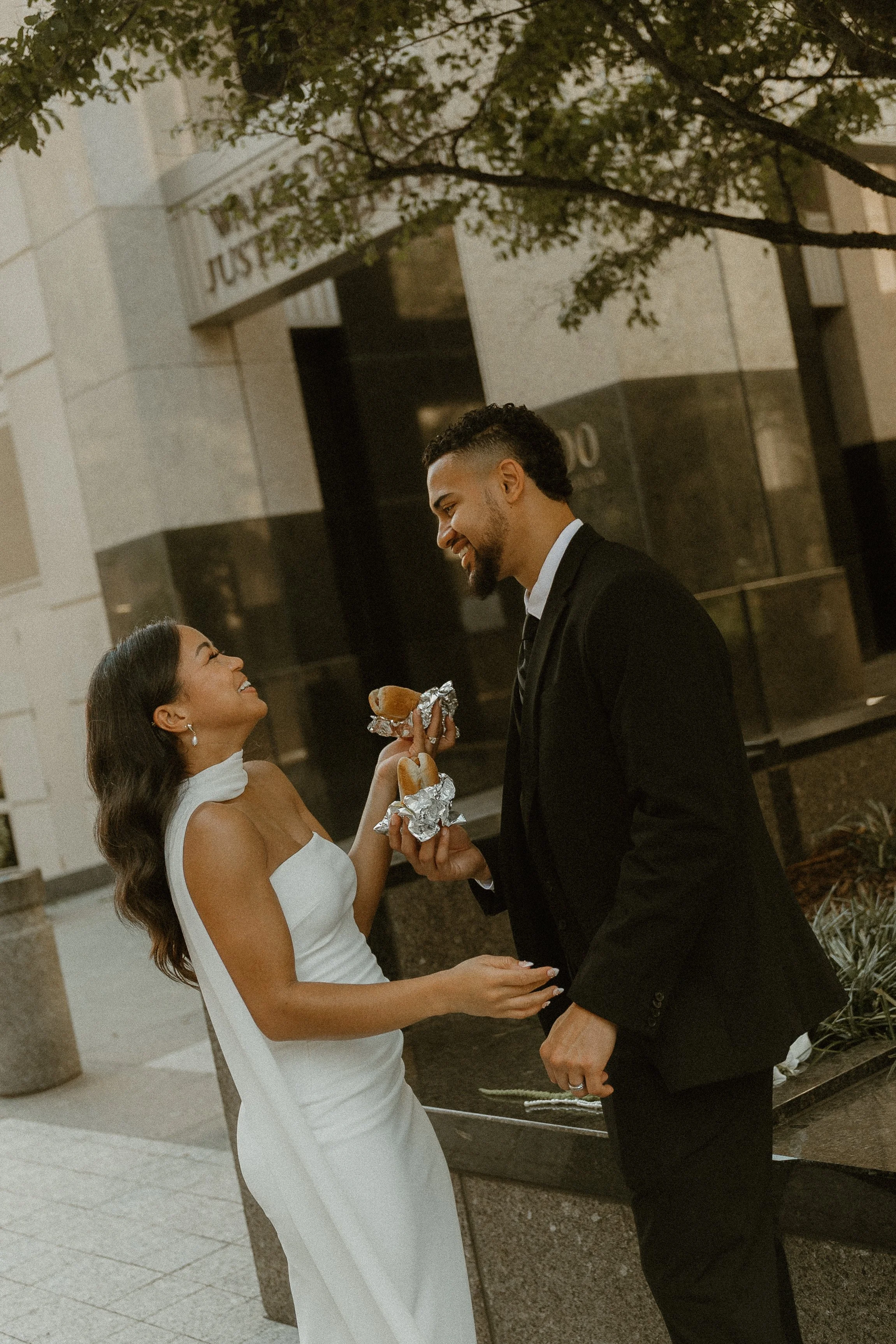 A couple dressed in formal attire sharing a moment outdoors, with the woman giving a piece of bread to the man, both smiling and looking at each other.