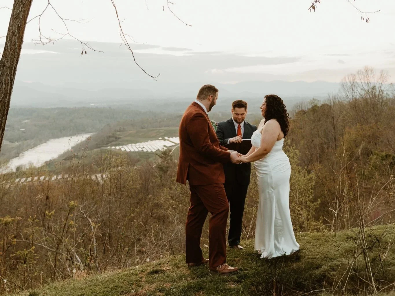 A couple dressed in wedding attire standing in a cozy room, holding hands and gazing at each other, with large windows and decorative interior elements including a couch, art on the wall, and a table with flowers.