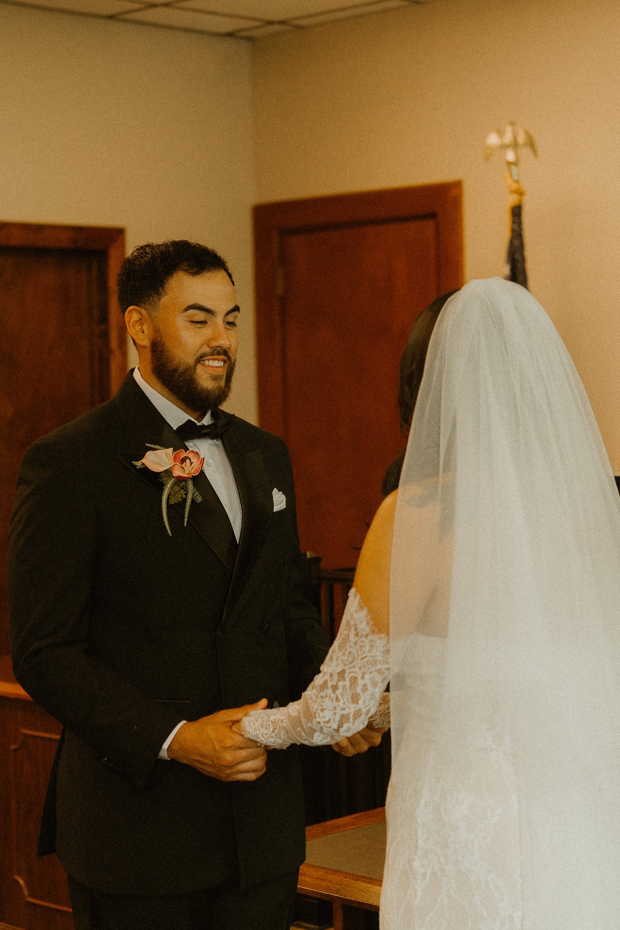 A groom in a black tuxedo with a pink boutonniere and a groomer-style beard is holding hands with a bride who is wearing a white lace wedding dress and veil. They are standing inside a room with wooden doors and beige walls, and the groom is smiling 