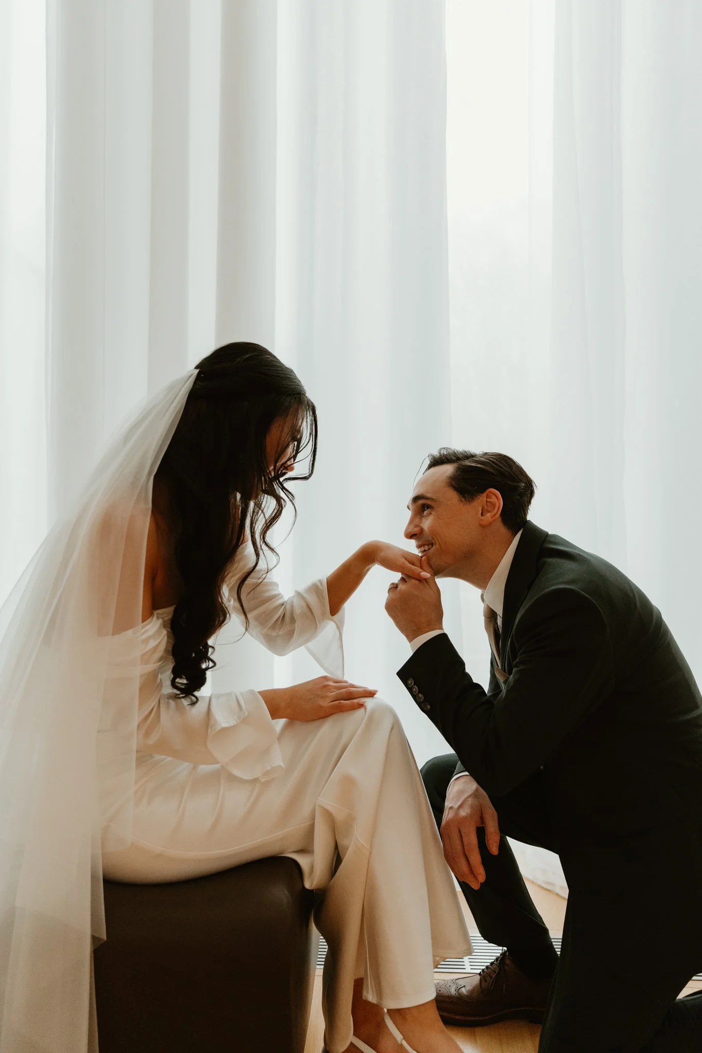 A bride in a white wedding dress and veil and a groom in a black suit, sitting near a window with white curtains. The groom is kissing the bride's hand while she looks down at him, with a warm and loving expression.