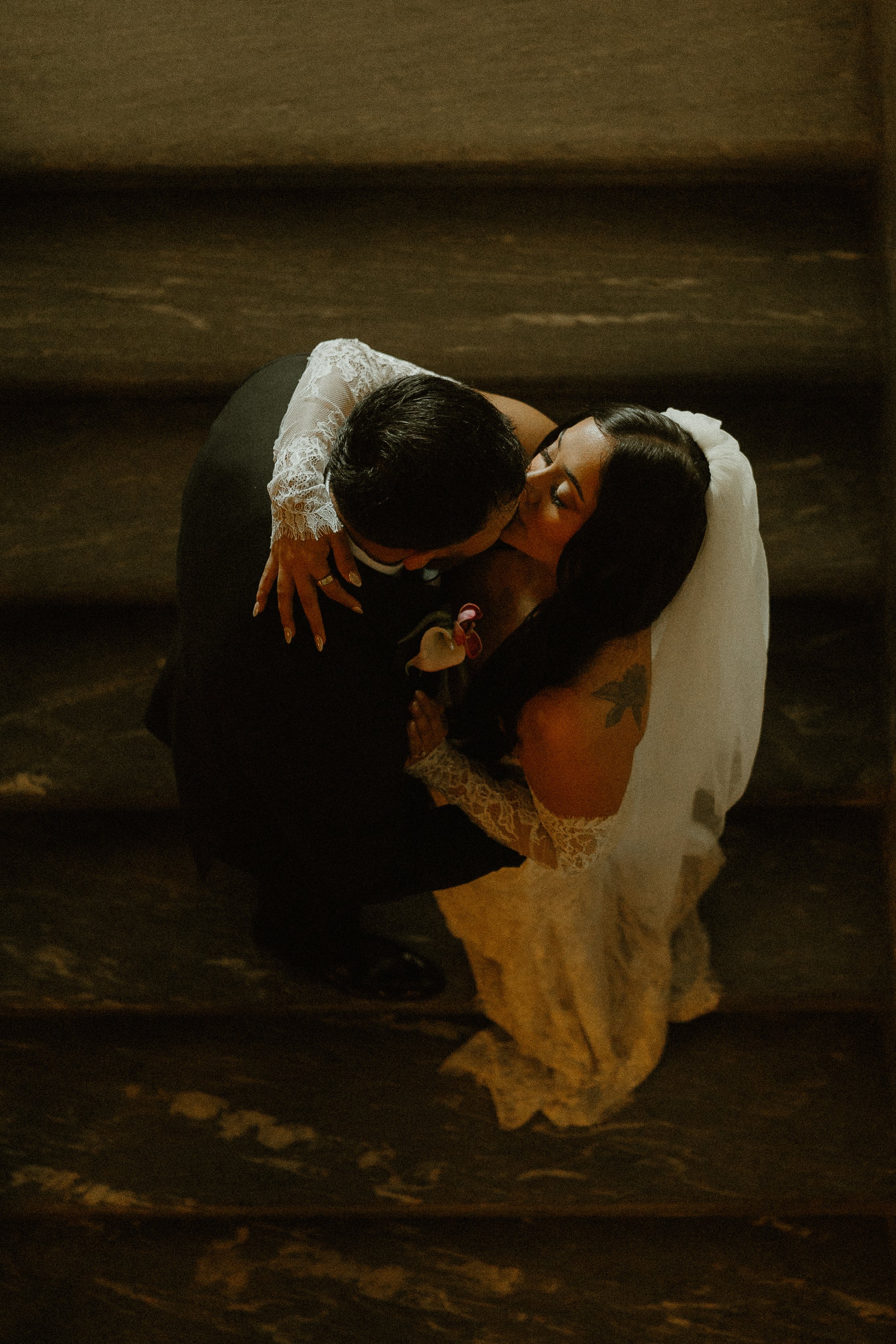 A couple sharing a kiss on their wedding day, with the bride wearing a white lace dress and veil, and the groom in a dark suit, embracing in a dimly lit setting.