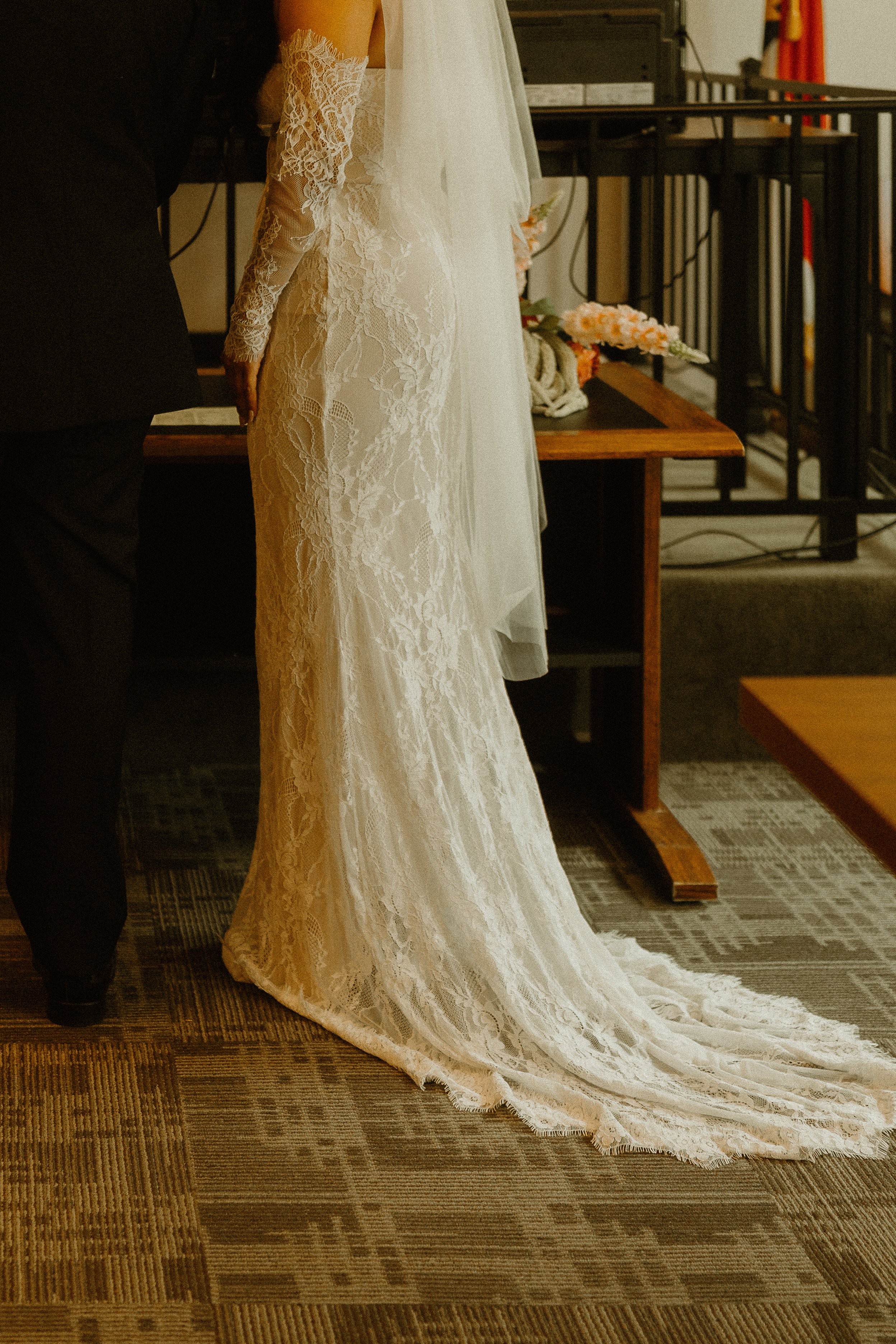 Close-up of a bride's long lace wedding dress train, with a person assisting her, standing on a patterned carpet in an indoor setting with wooden furniture and a black railing.