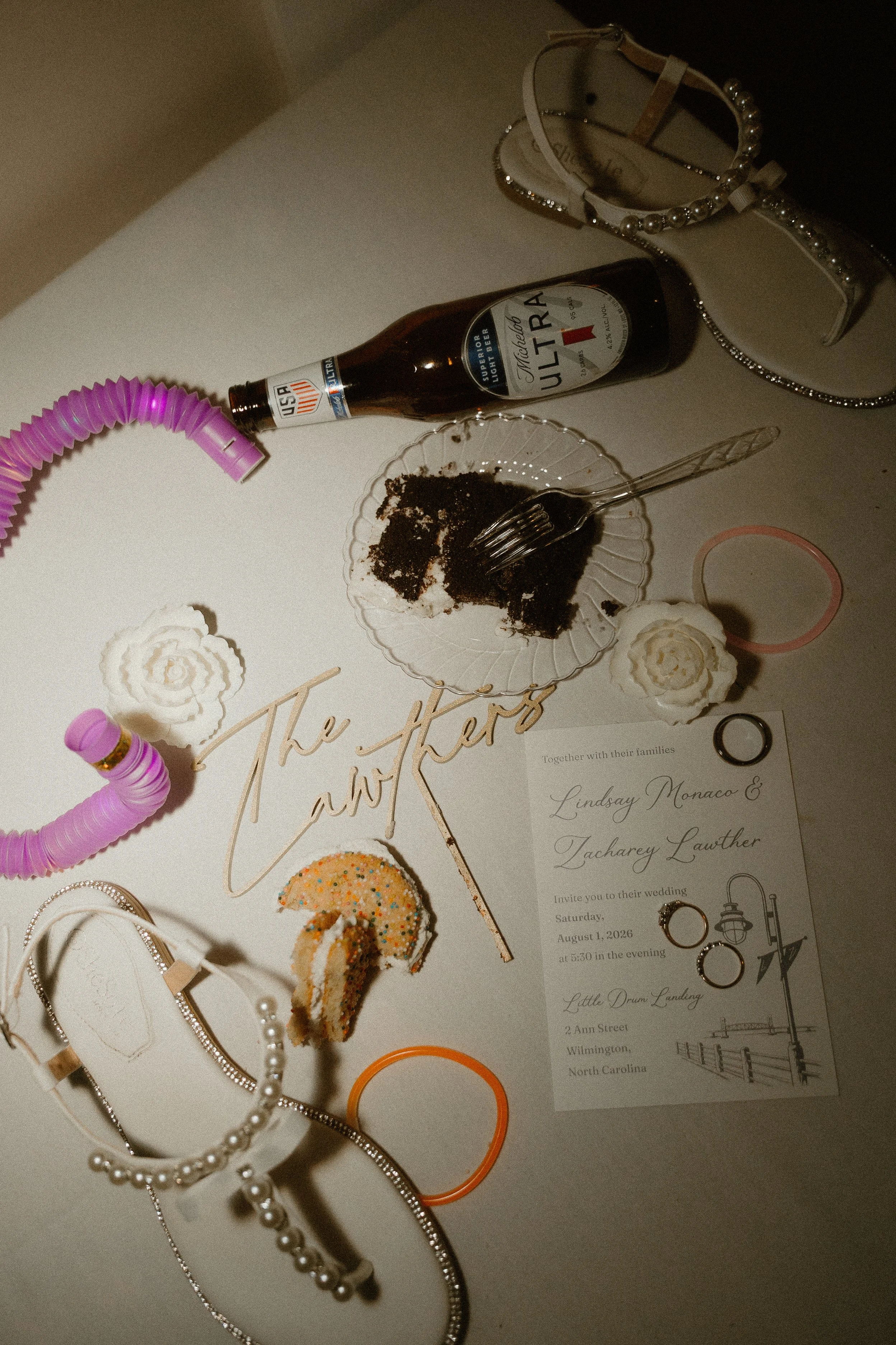 A table setup showing a Michelob Ultra beer bottle, a slice of chocolate cake on a glass plate with a fork, decorated cookies, various jewelry including necklaces, rings, and hairbands, and an wedding invitation for Lindsay Monaco and Tacharey Lawthe