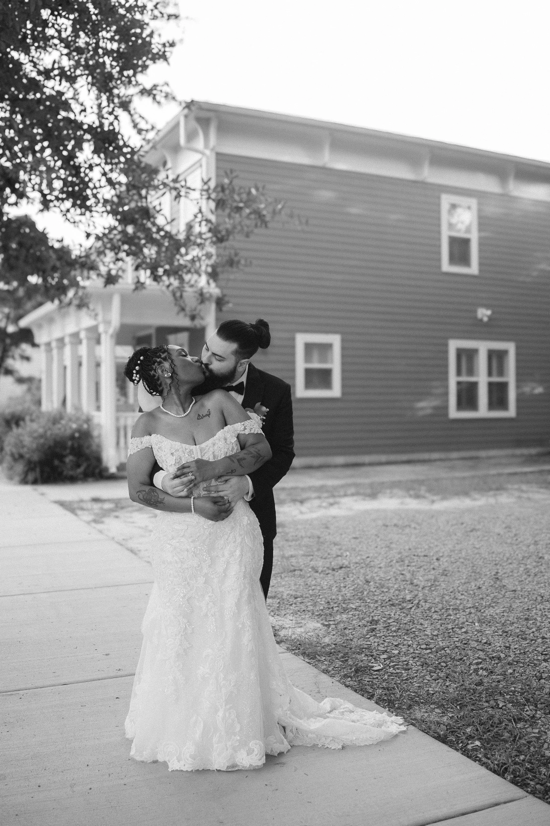 A bride and groom sharing a kiss outdoors in front of a house, with the bride in a white lace wedding gown and the groom in a black tuxedo.