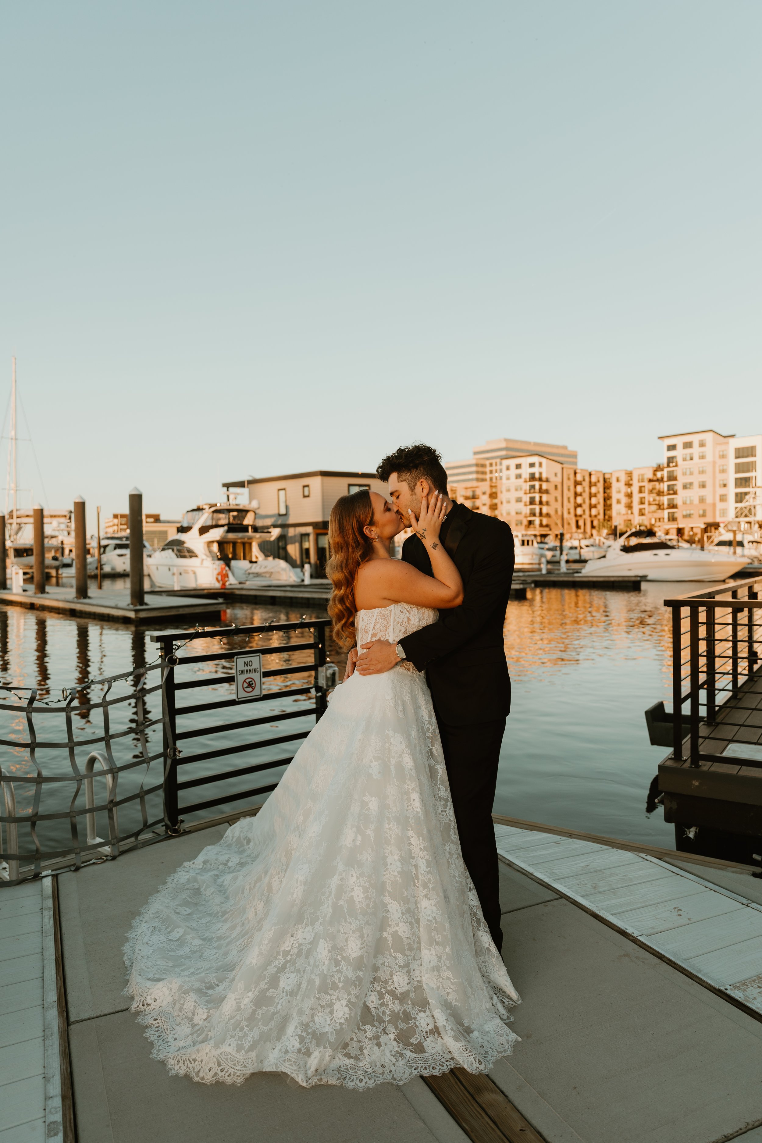 A bride and groom kissing on a dock at a marina during sunset, with boats and apartment buildings in the background.