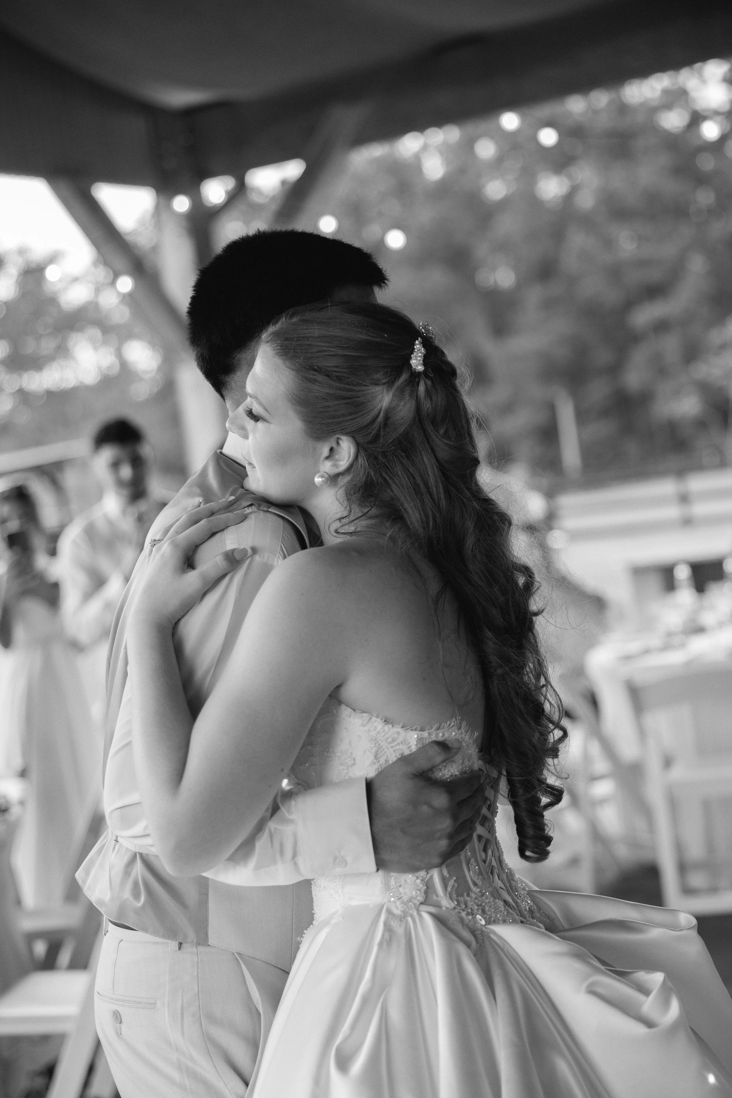 A bride and groom passionately dance and embrace each other at a wedding reception, with guests in the background. The bride has long hair with a decorative hairpiece and wears a strapless wedding gown. The groom wears a light-colored suit with a dre