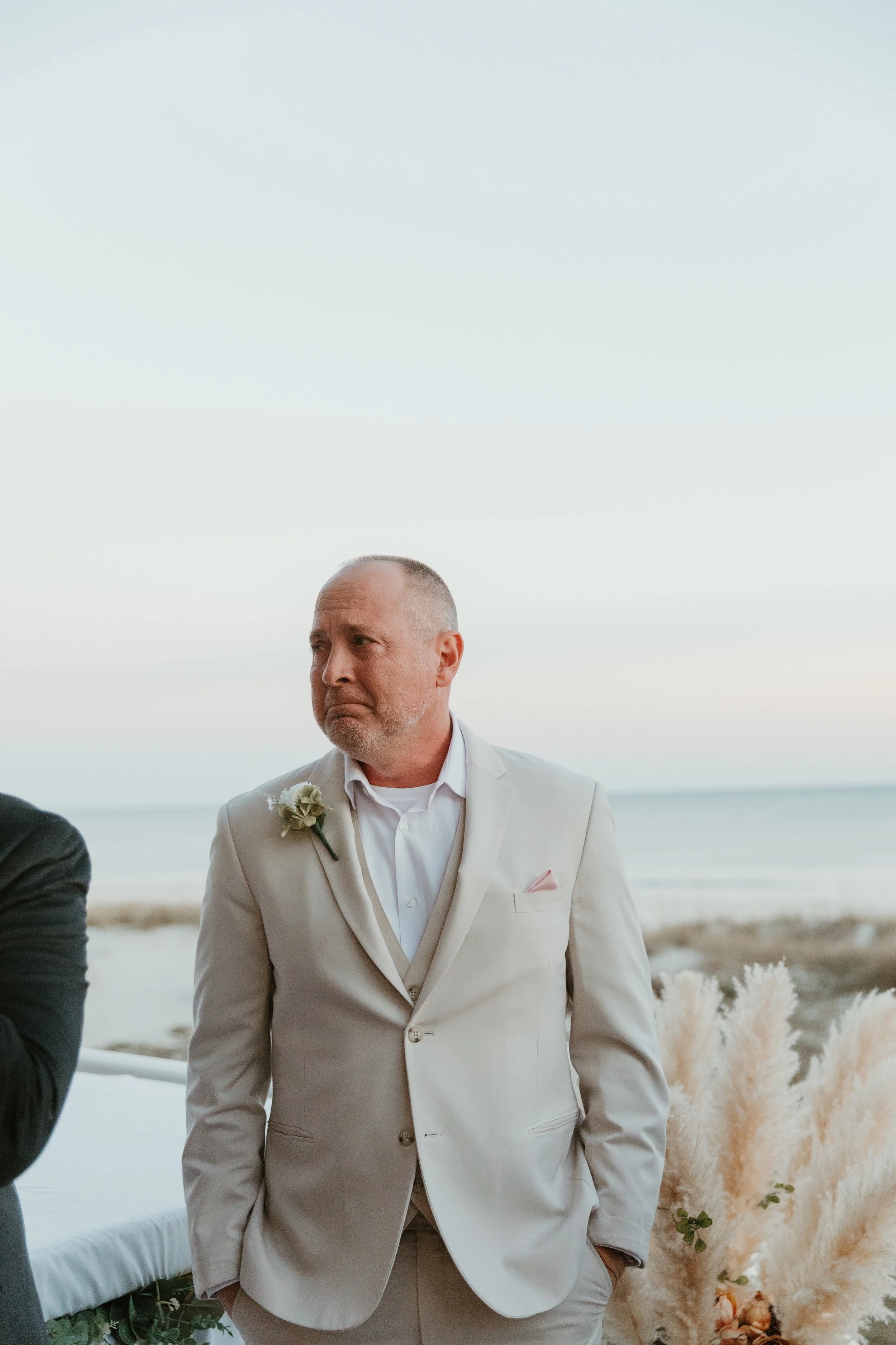A man in a white suit at a beach wedding, standing with his hands in his pockets, looking to the side.