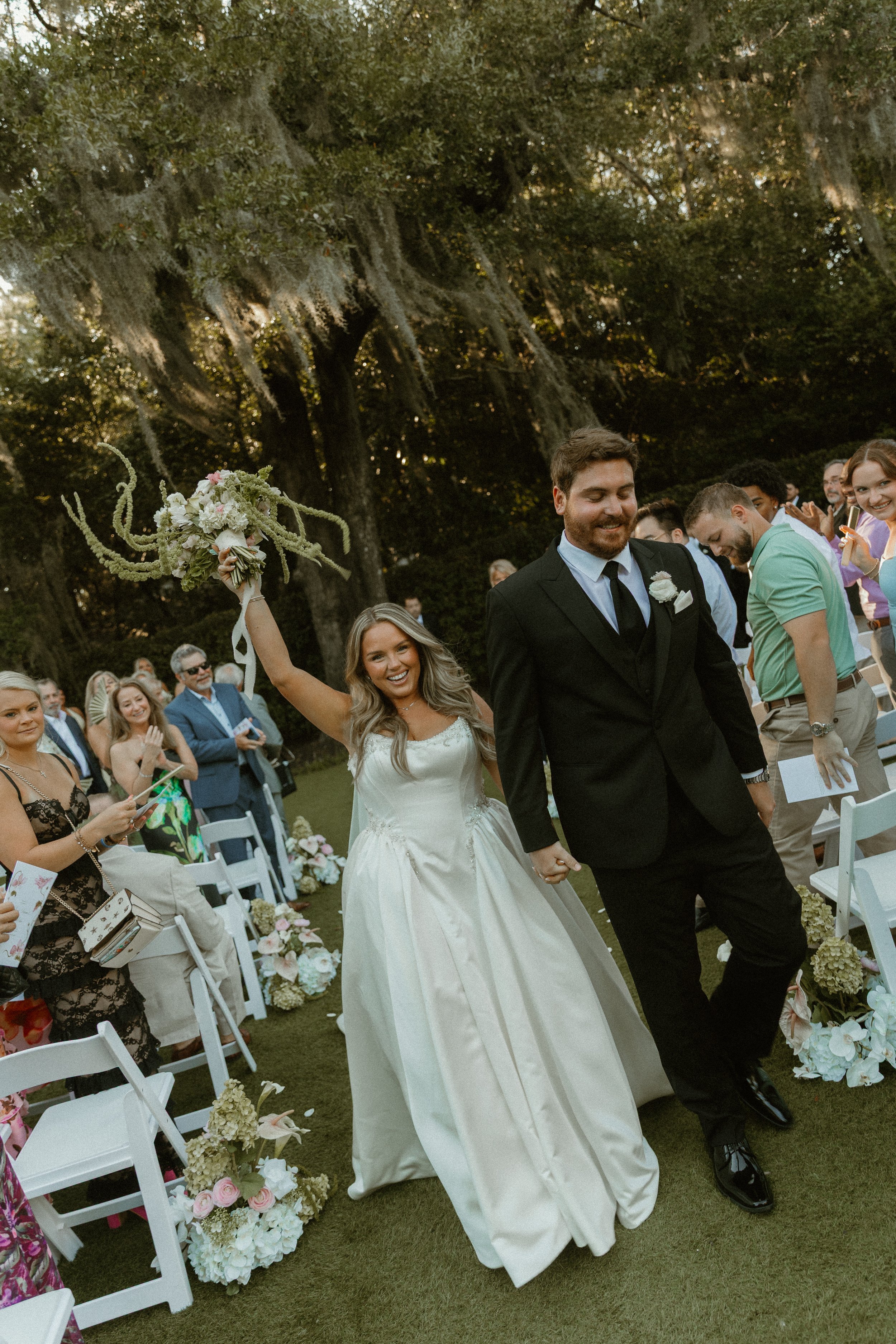 A bride and groom holding hands and walking down the aisle during their outdoor wedding ceremony, surrounded by seated guests and floral arrangements, with large trees in the background.