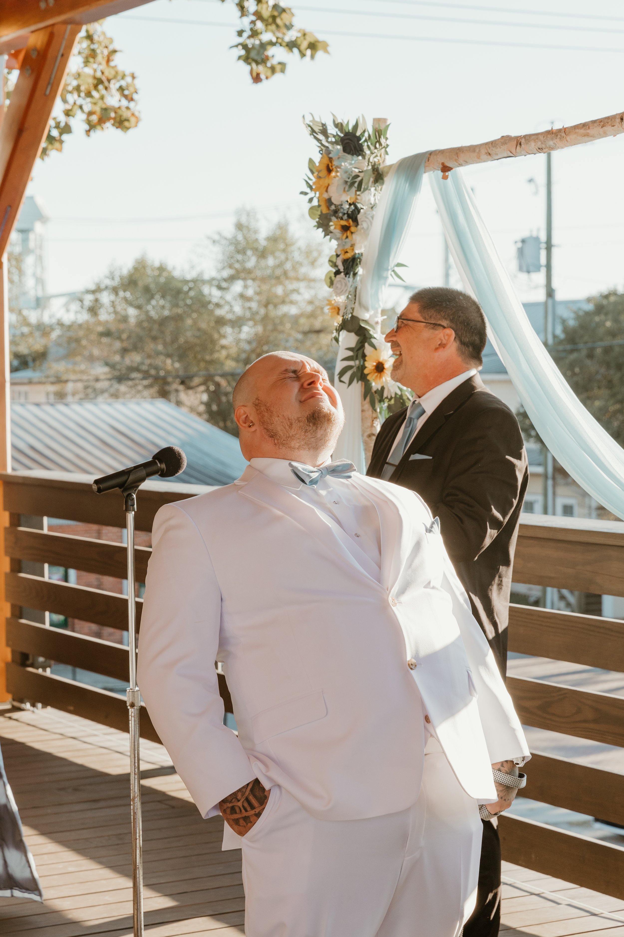Groom in a white tuxedo, with tattoos visible on his hand, is emotional during his wedding ceremony, standing on an outdoor wooden platform with an officiant in a black suit behind him, in front of a floral wedding arch with fabric decorations.