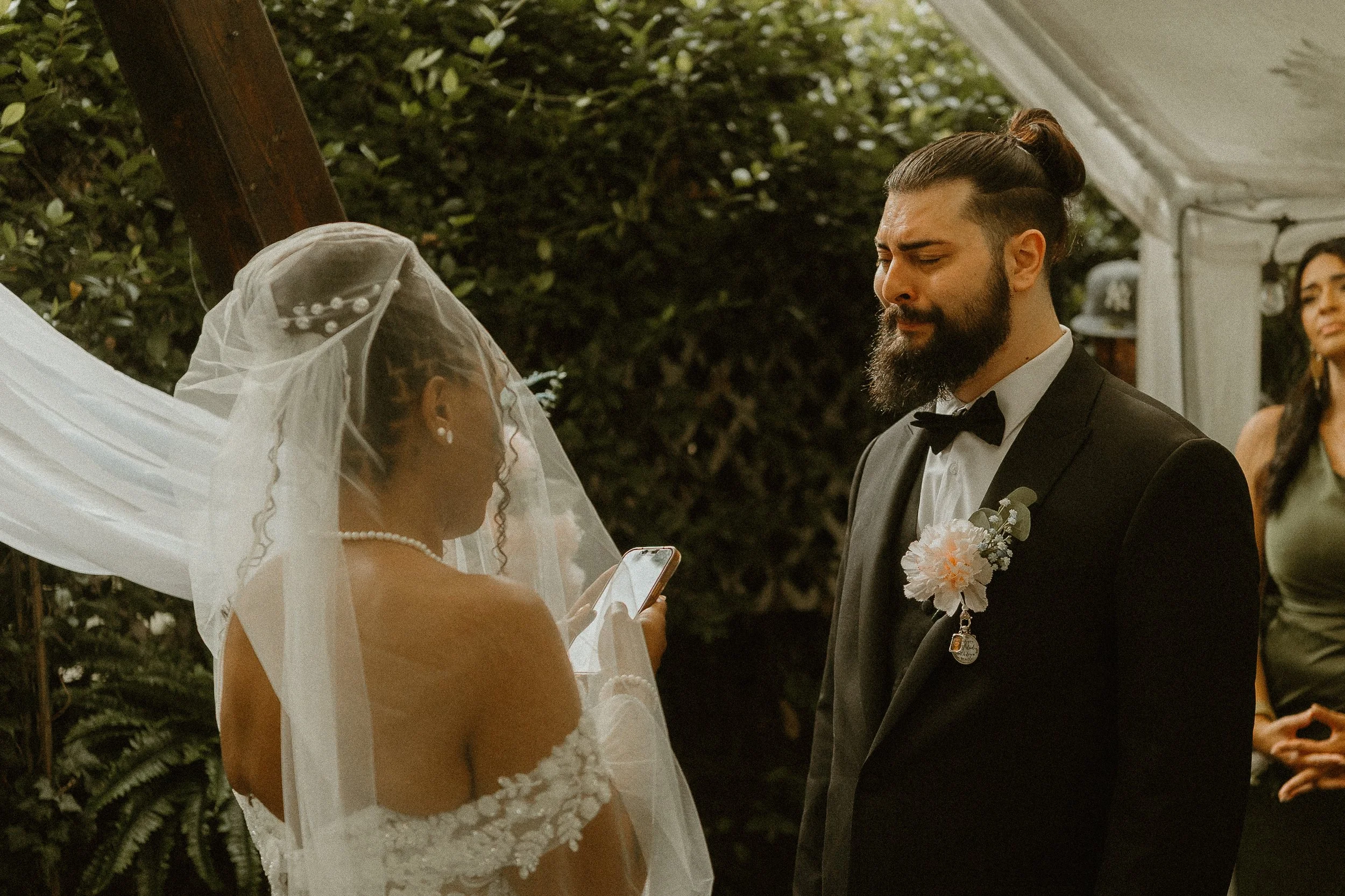 A wedding ceremony with a bride in a lace wedding dress and veil reading vows from a phone, and a groom in a black tuxedo with a boutonniere and bow tie, standing opposite each other outdoors surrounded by greenery.