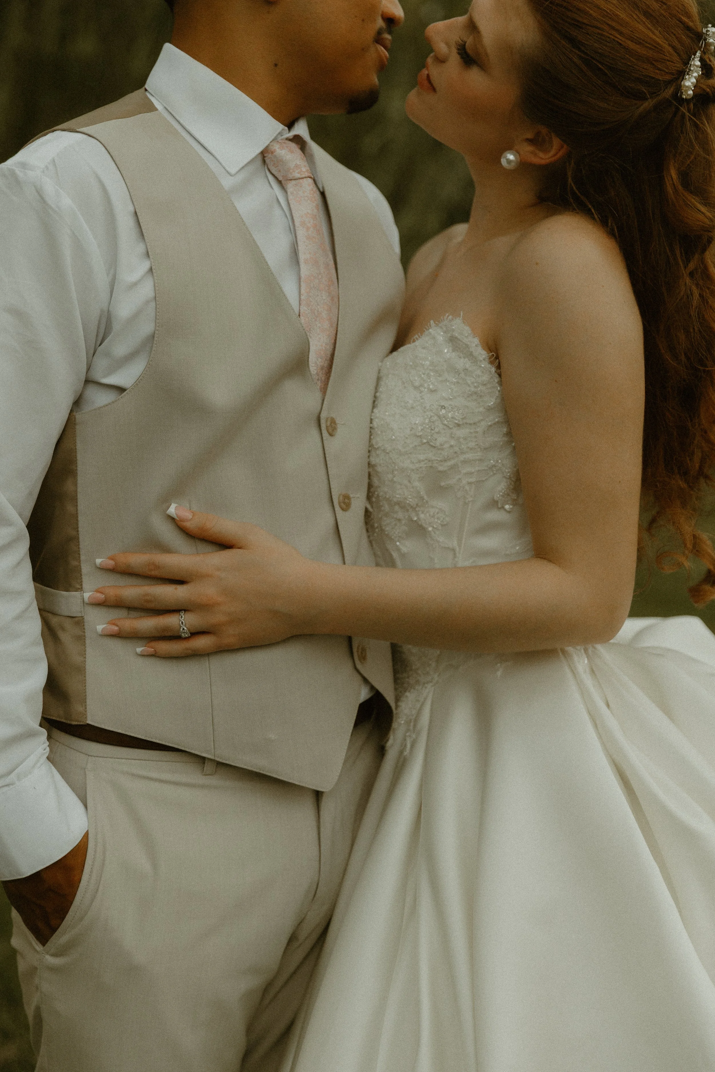 A bride and groom close together, about to kiss, in wedding attire. The groom wears a beige vest and pants with a white shirt and pink tie. The bride wears a white strapless wedding dress with lace details, pearl earrings, and has red hair styled wit