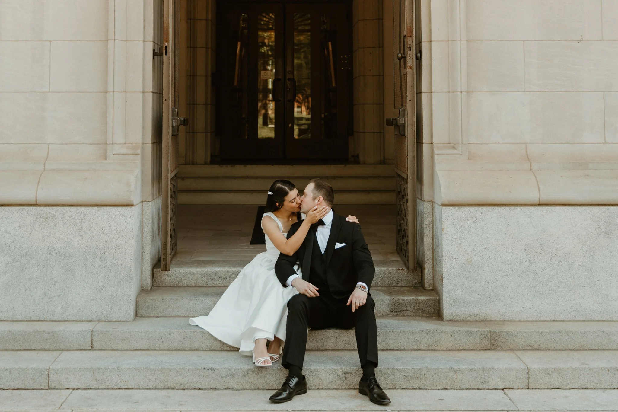 A bride and groom sitting on the steps of a building, sharing a kiss, with the bride wearing a white wedding dress and the groom in a black tuxedo.