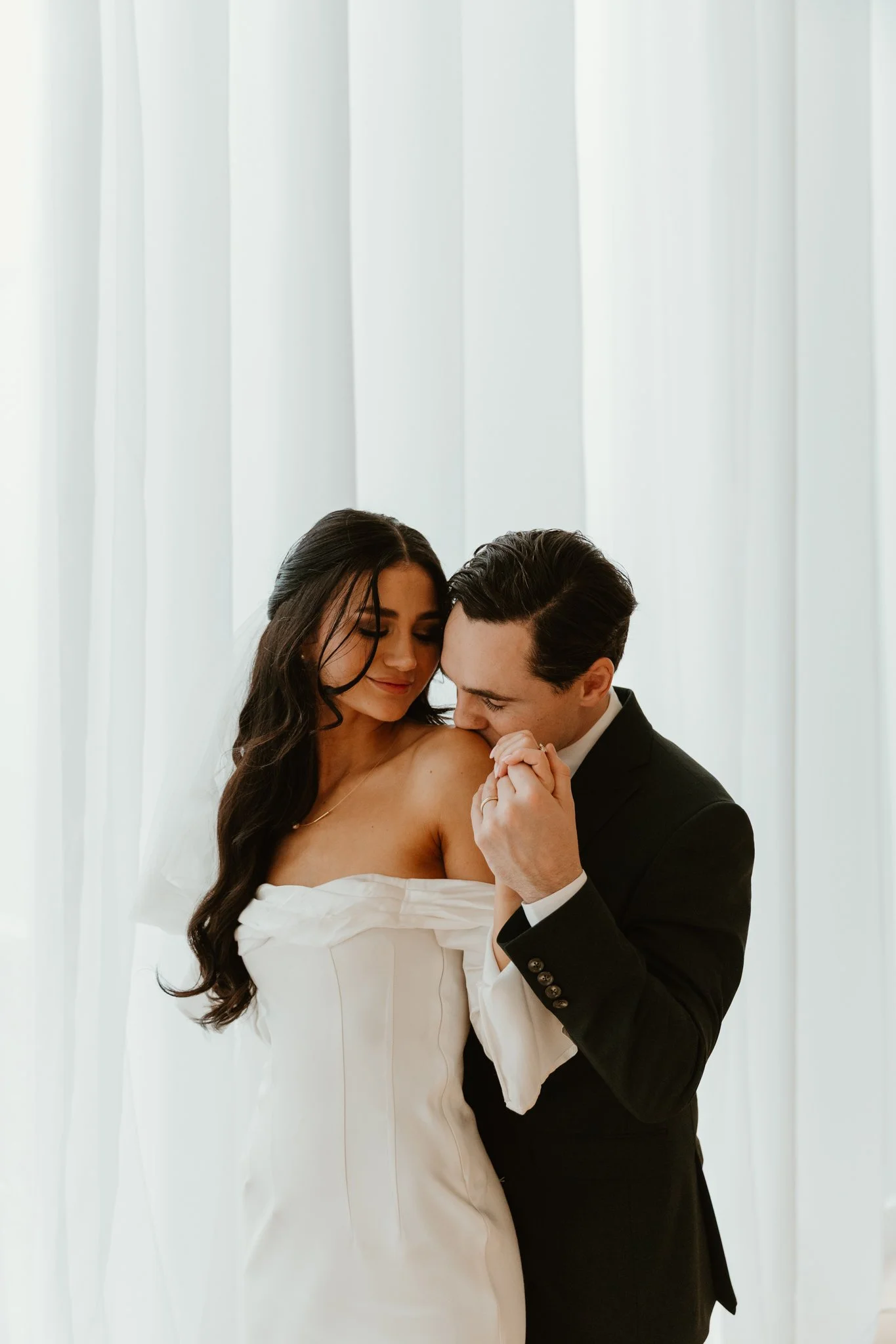 A bride and groom sharing a tender moment indoors, with white curtains in the background.