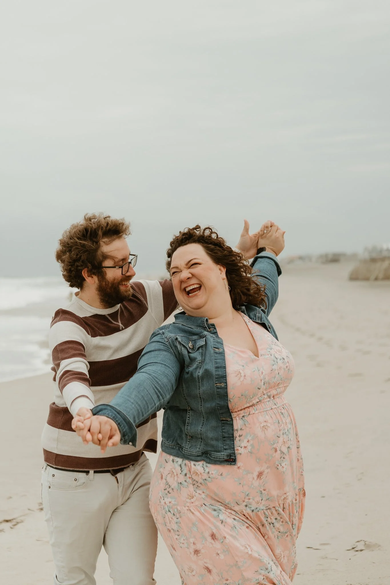 A group of four friends holding hands and dancing on a beach with gray skies, smiling and enjoying themselves.