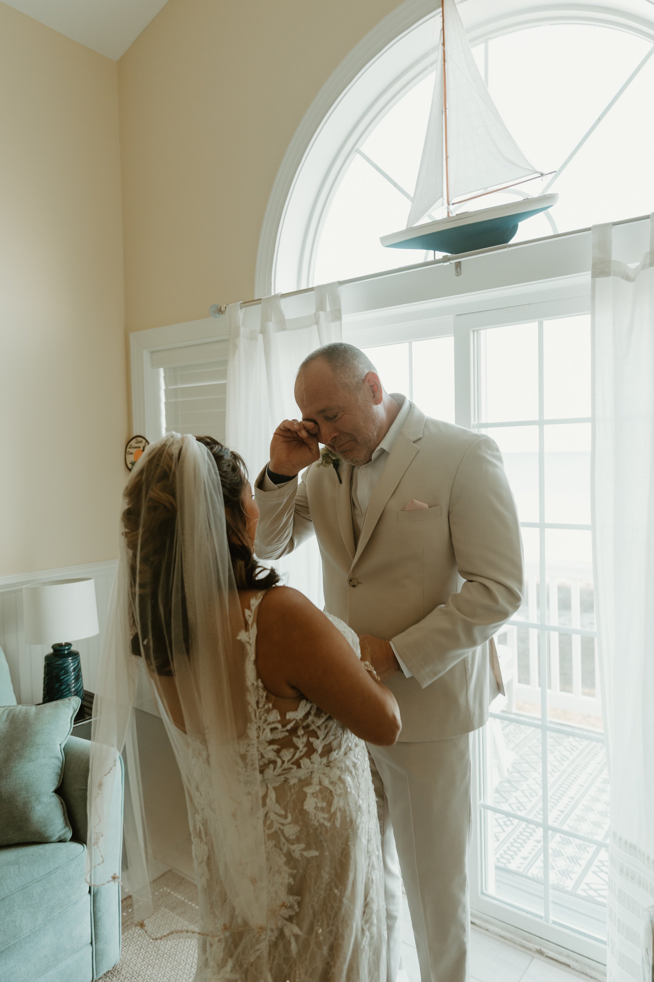 A groom in a white suit comforts a bride in a lace wedding dress and veil inside a sunny room with large windows and a model sailboat on the window ledge.