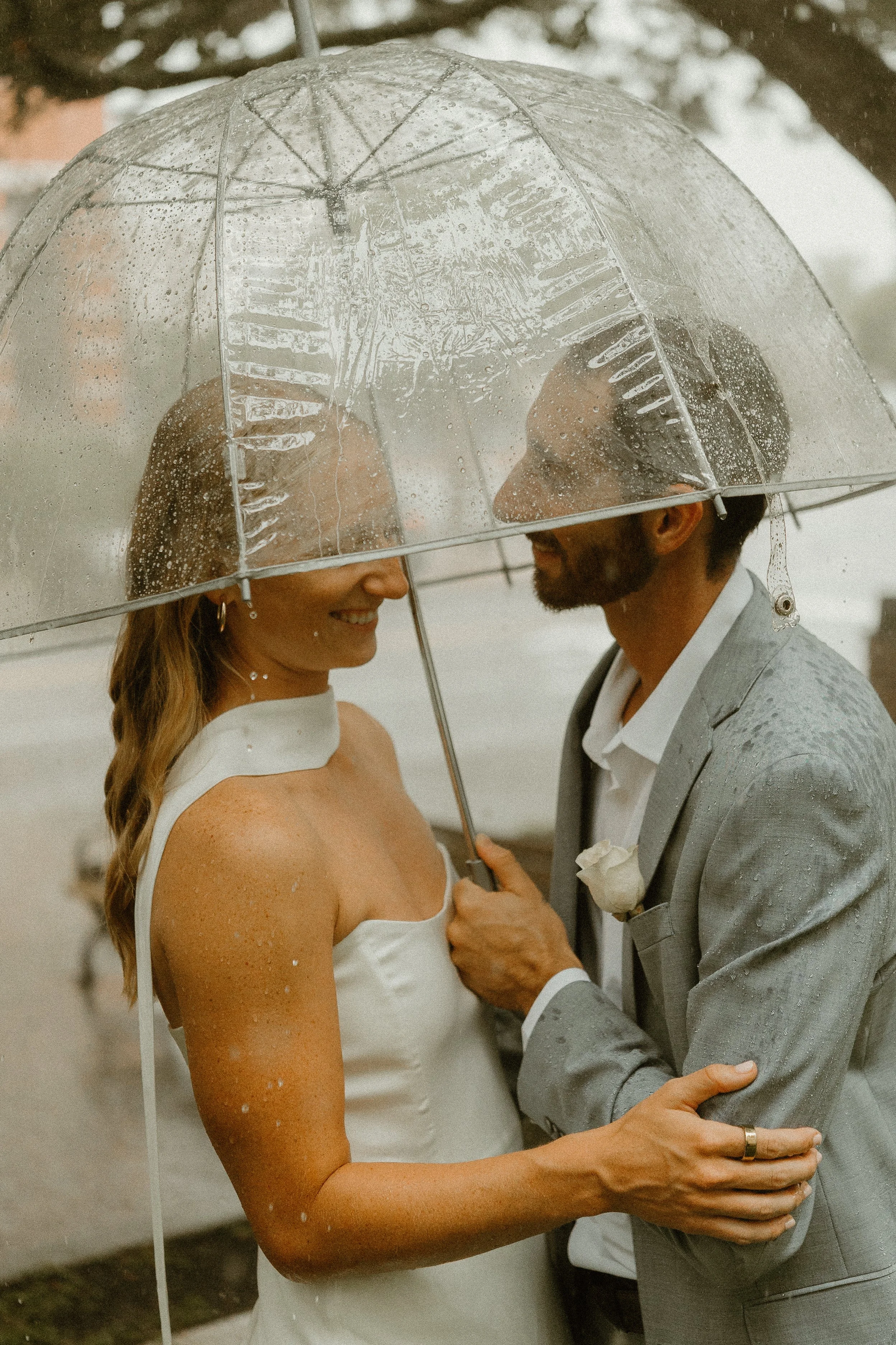 A couple standing close together under a transparent umbrella on a rainy day, smiling at each other. The woman is wearing a white dress with small embellishments, and the man is dressed in a light gray suit with a white shirt and a boutonniere. They 