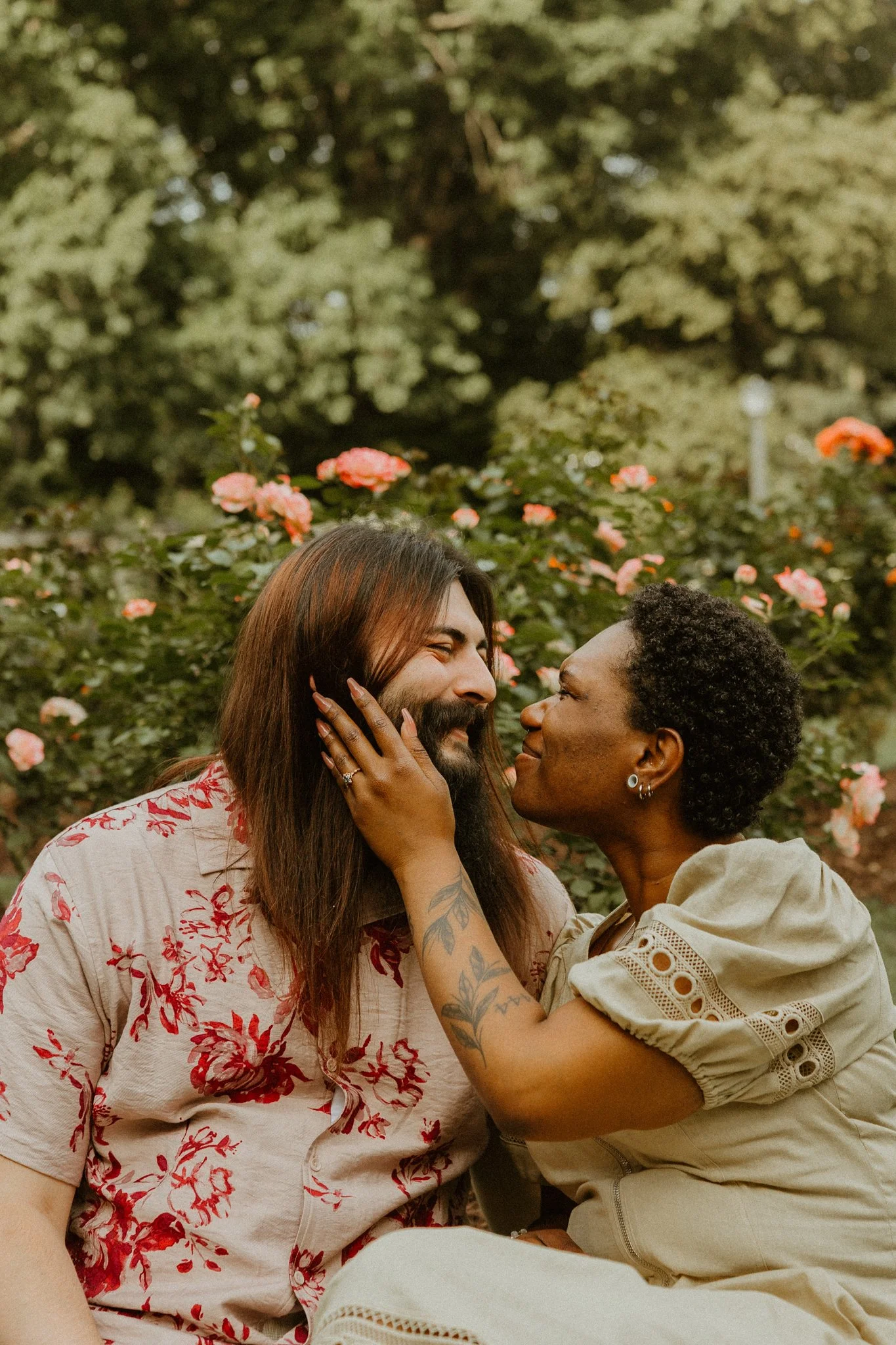 Two women, one with long brown hair and tattoos, and the other with short curly hair, are close together outdoors, touching faces, with a background of pink flowers and green foliage.