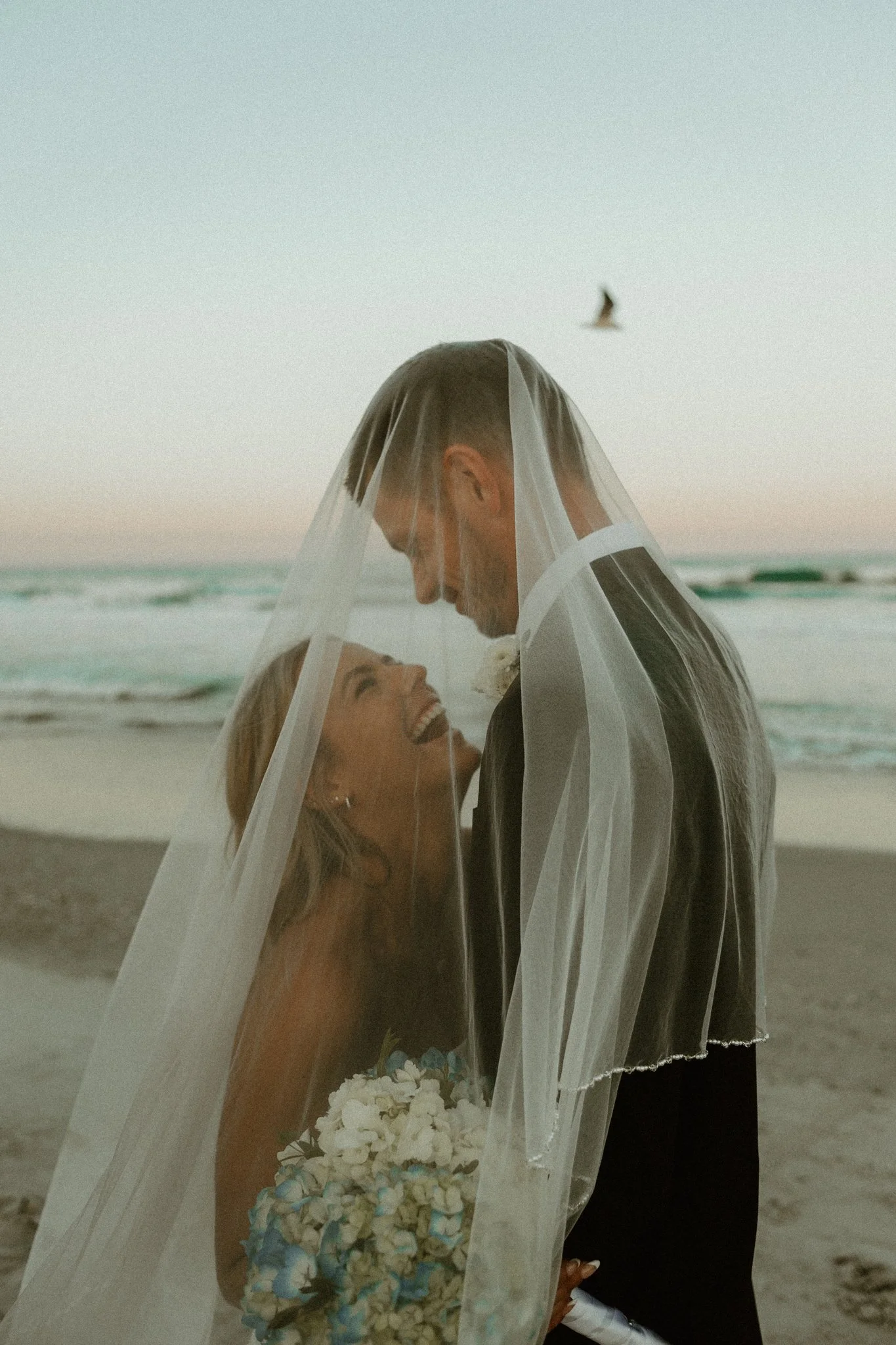 A couple at the beach couples in wedding attire, with the bride wearing a veil and holding a bouquet, and the groom in a suit. They are smiling and looking at each other, with a bird flying in the background.