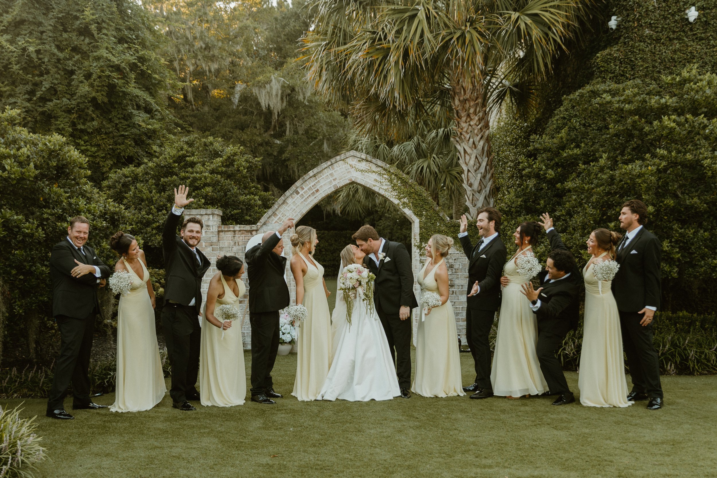 A group of wedding guests in formal attire celebrating outdoors, with a bride and groom kissing in the center, surrounded by lush greenery, palm trees, and a stone archway.