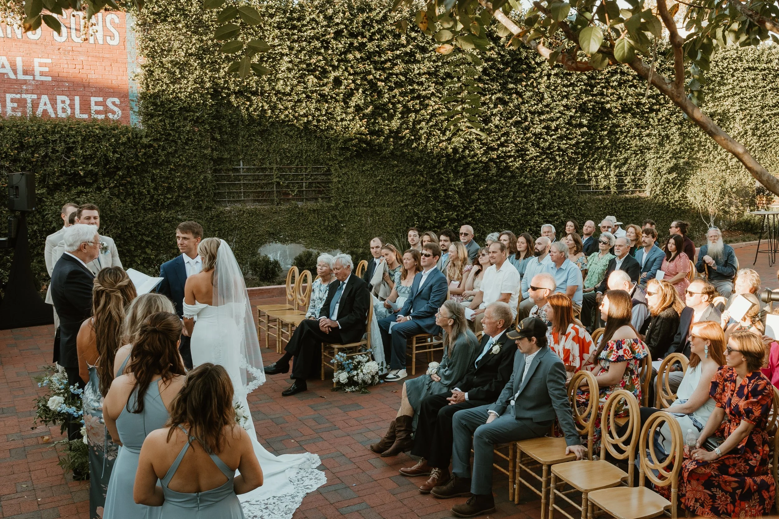 Outdoor wedding ceremony with guests seated on wooden chairs on brick pavement, facing an officiant and a bride and groom standing under a large tree. Greenery and flowering bushes surround the scene. A large sign on a brick wall partially in view reads 'YOUR SALE VEGETABLES.'