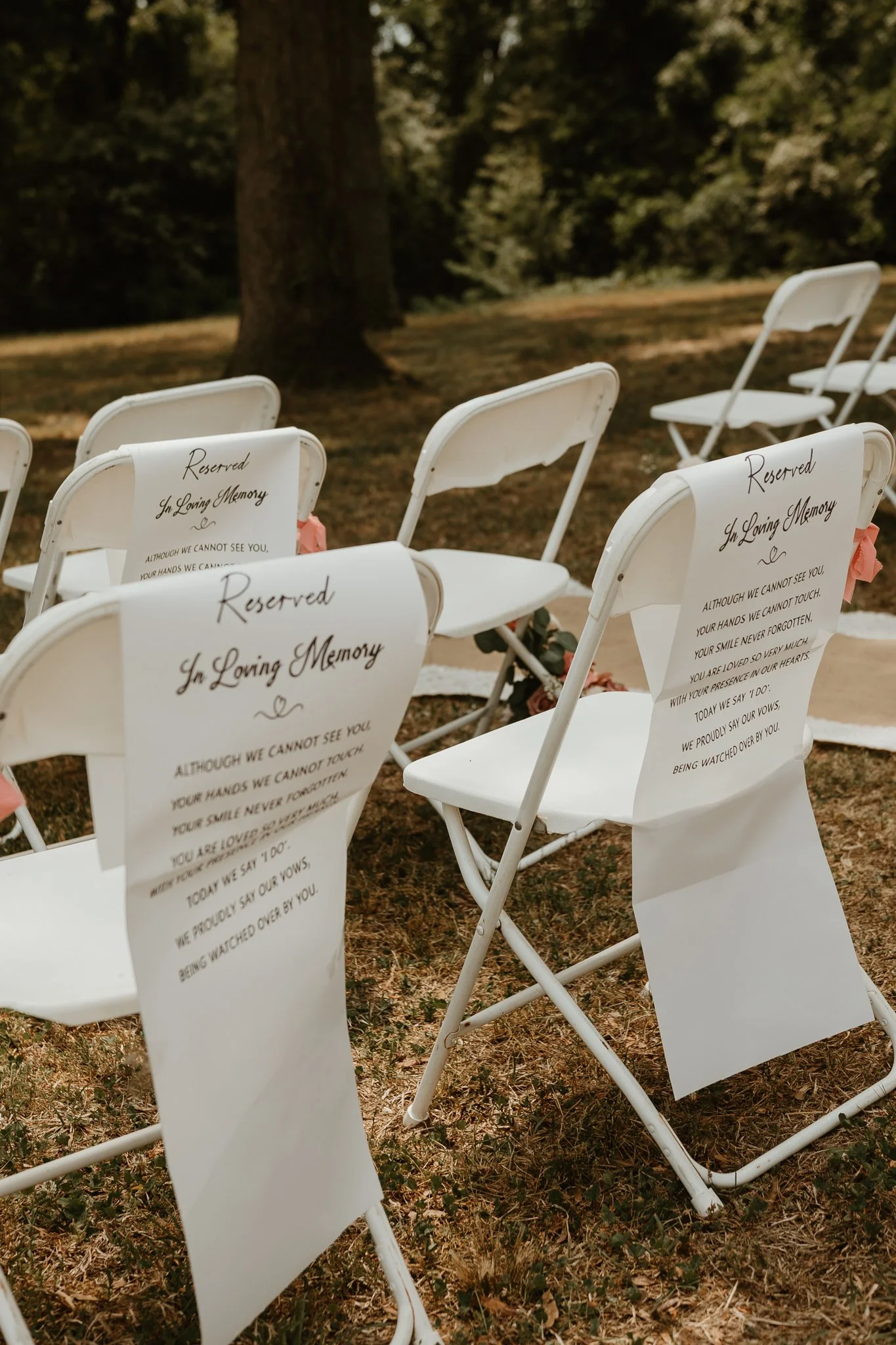 Row of white chairs with reserved signs in memory of loved ones, outdoors on grass with trees in background.