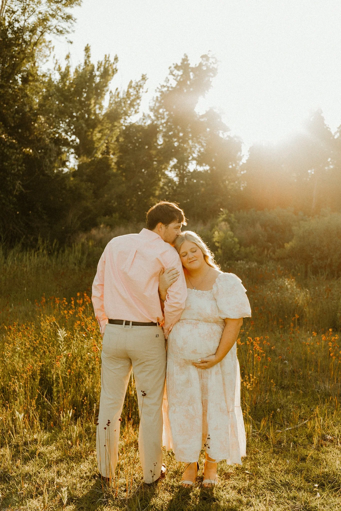 A couple stands in a field of wildflowers at sunset, with trees in the background. The woman is pregnant, and the man is resting his head on her shoulder.