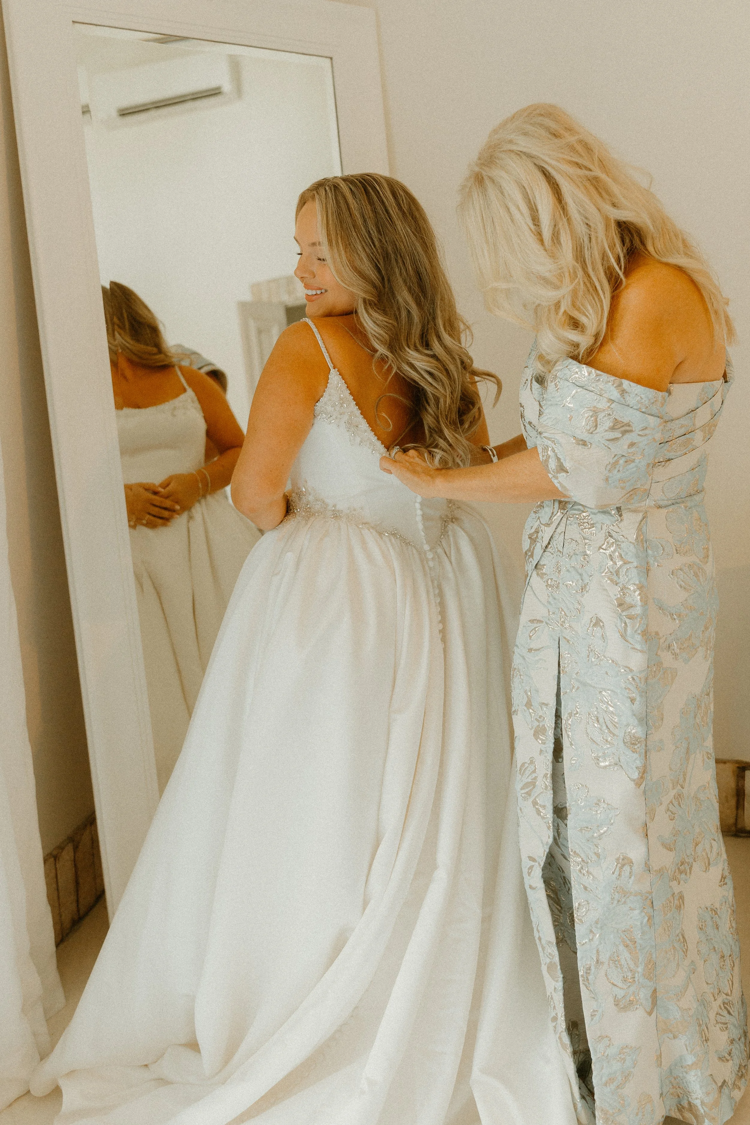 A woman in a wedding dress is smiling in front of a mirror as another woman helps her adjust it. The bride has long, styled hair, and the women are in a bright room.