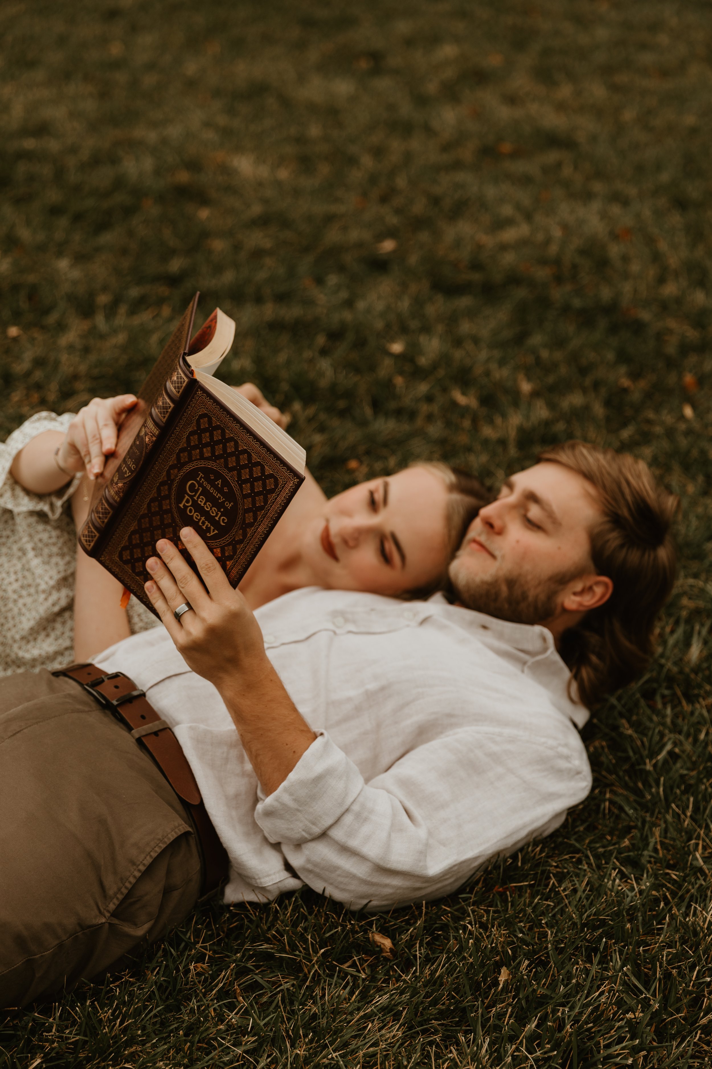 A couple lying on grass, the woman resting her head on the man's shoulder while reading a book titled 'Treasury of Classic Poetry'.
