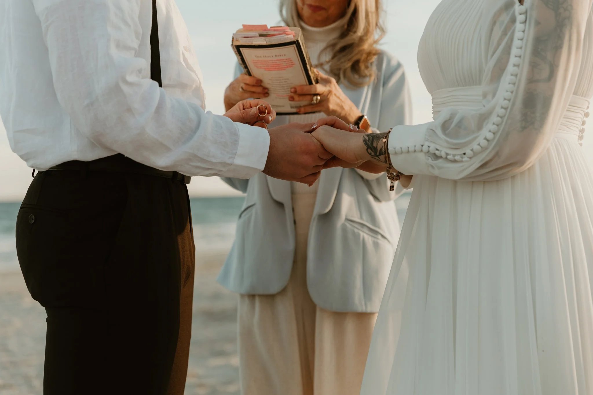 A couple holding hands during a wedding ceremony at the beach, with a woman officiant reading vows in the background.