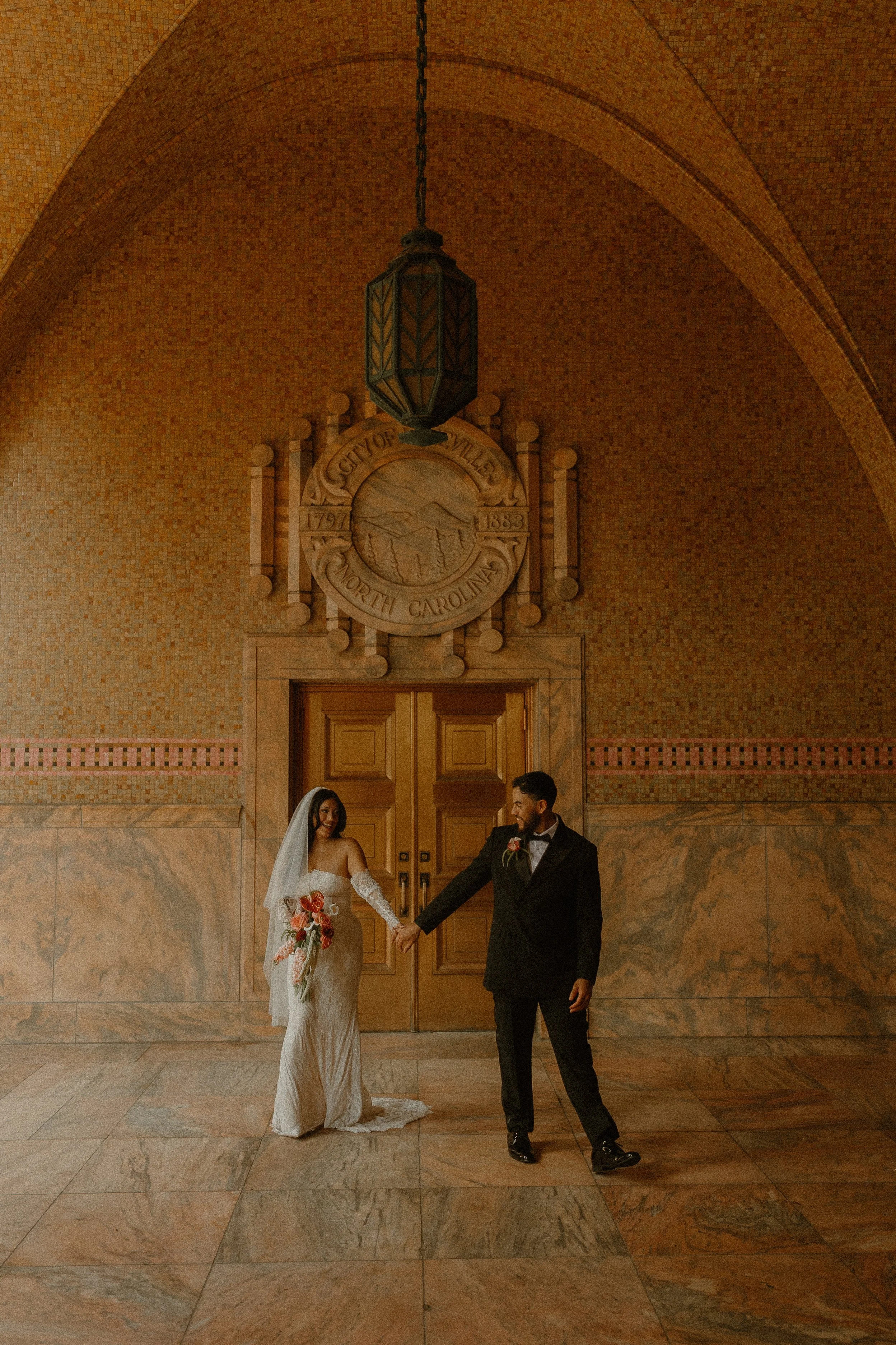 A bride and groom holding hands and smiling at each other are standing inside a grand, historic building with marble floors, an arched brick ceiling, and a carved emblem that reads 'City of Asheville North Carolina 1791-1883' behind them.