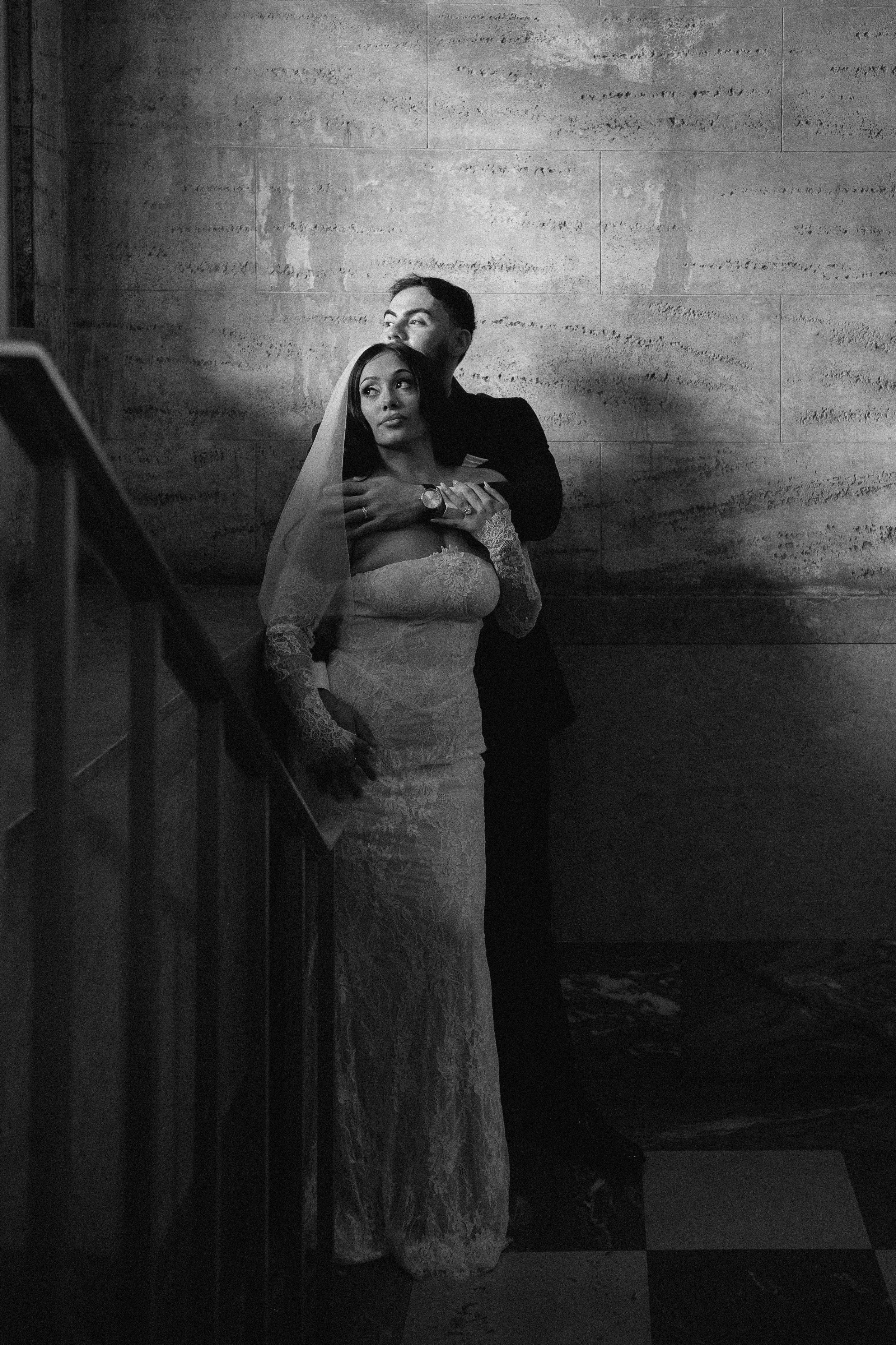 A black and white photo of a bride in a lace wedding dress and veil standing on a staircase, with a groom standing behind her, holding her, looking over her head, against a textured wall.