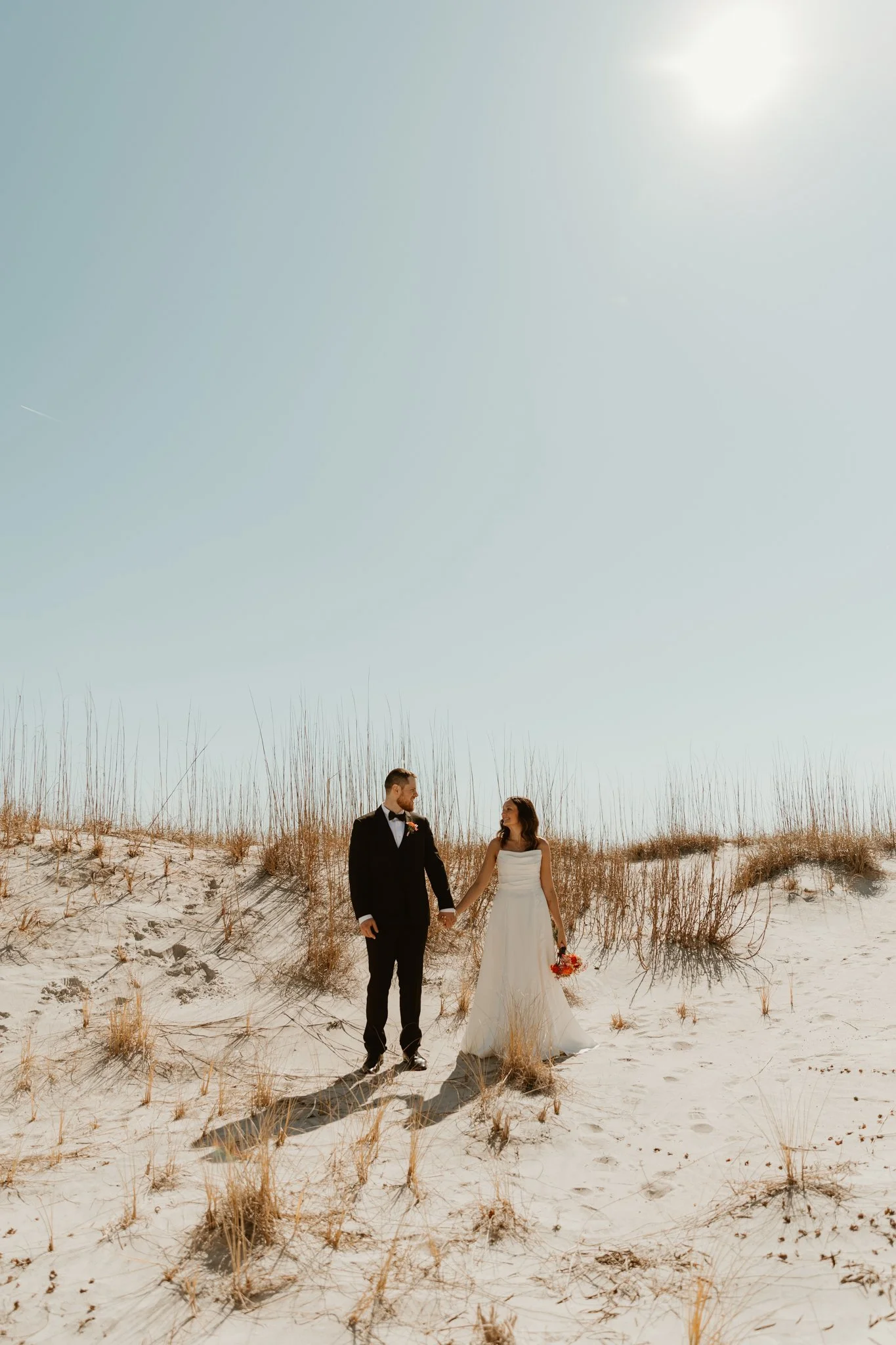 A bride and groom standing hand in hand on a sandy beach with tall grass, holding a bouquet, under a clear sky and bright sun.