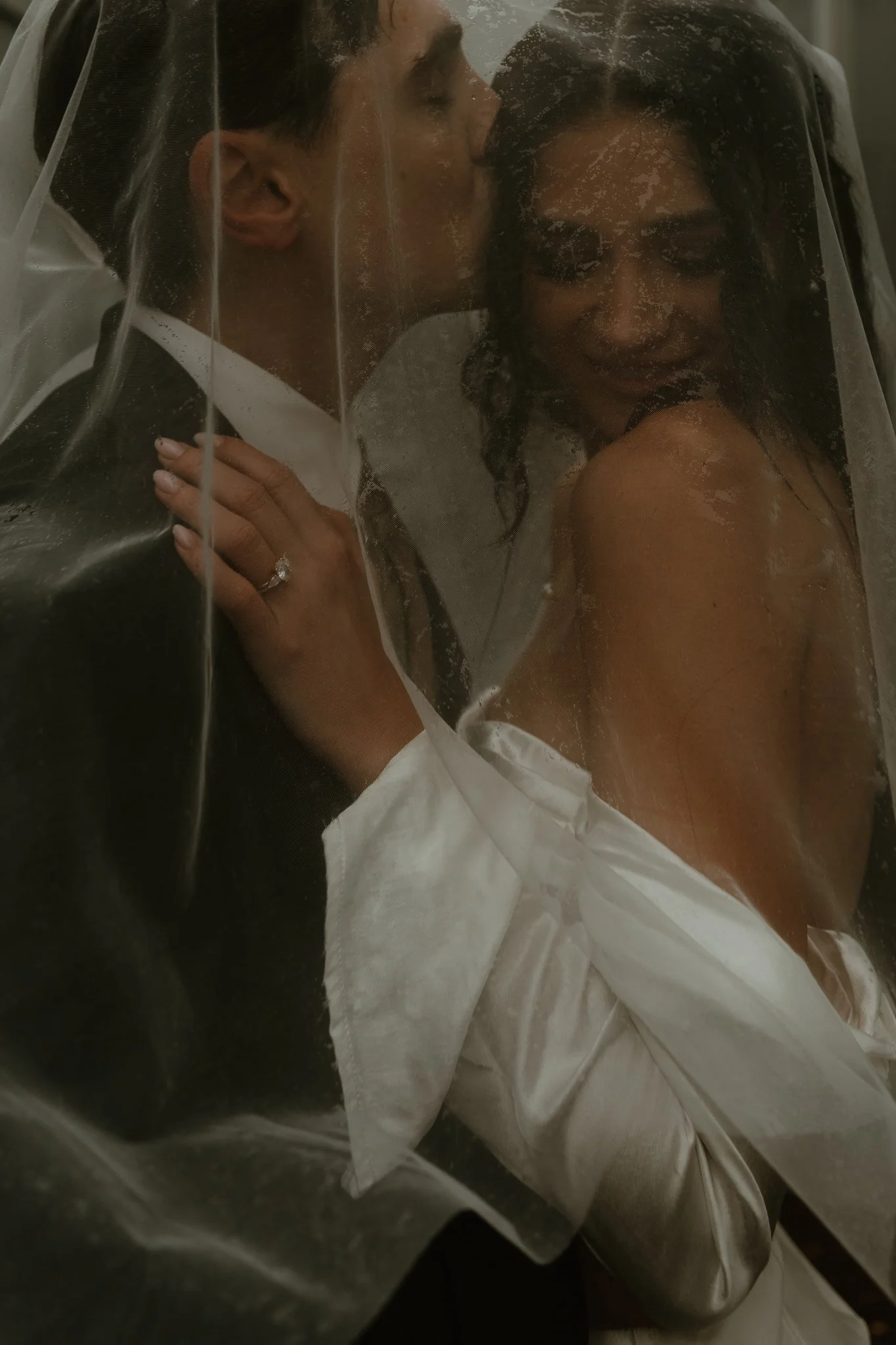 A bride and groom share an intimate moment behind a wedding veil, with the groom kissing the bride's forehead.