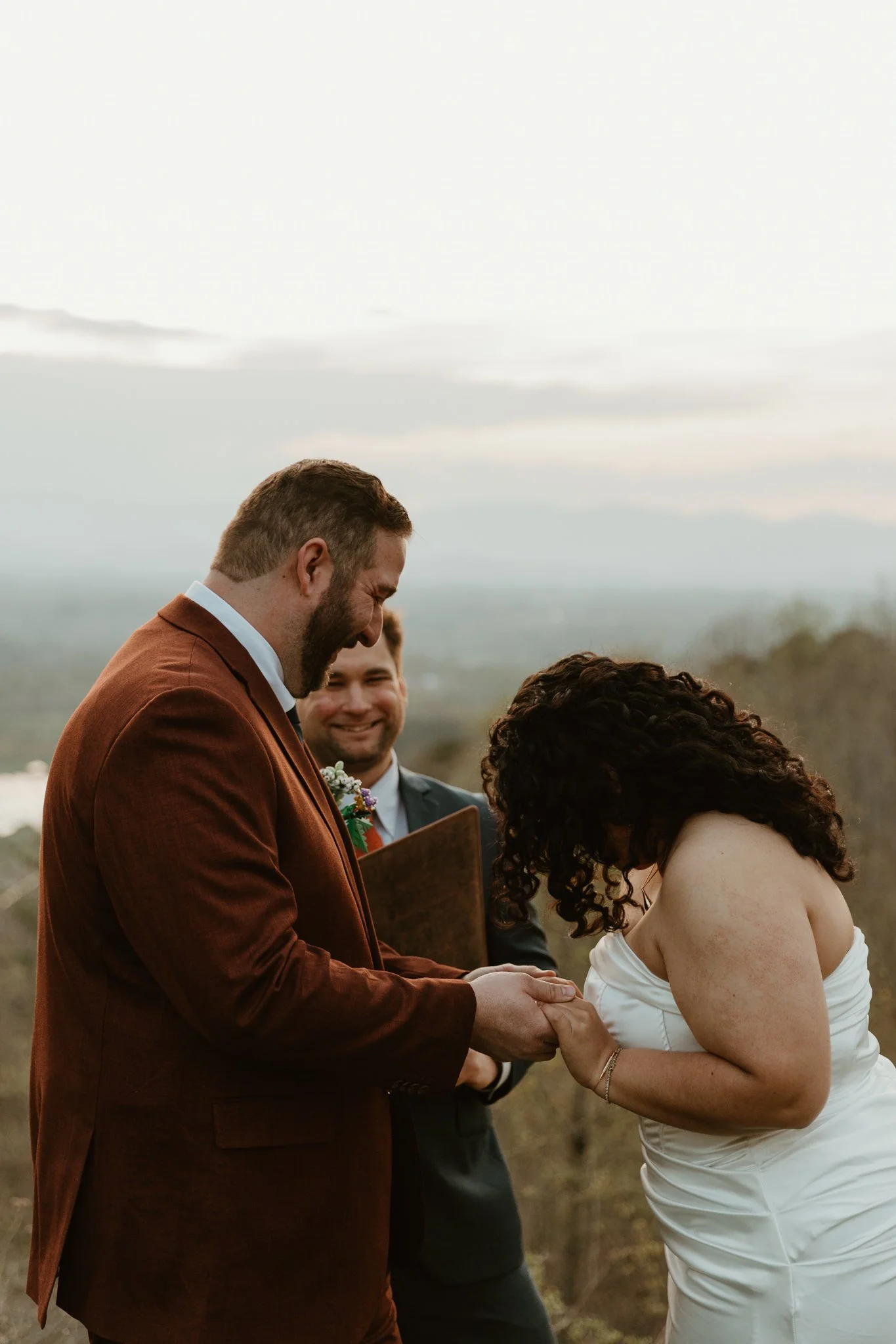 A bride and groom holding hands during their wedding ceremony outdoors, with a minister reciting vows and guests smiling in the background.