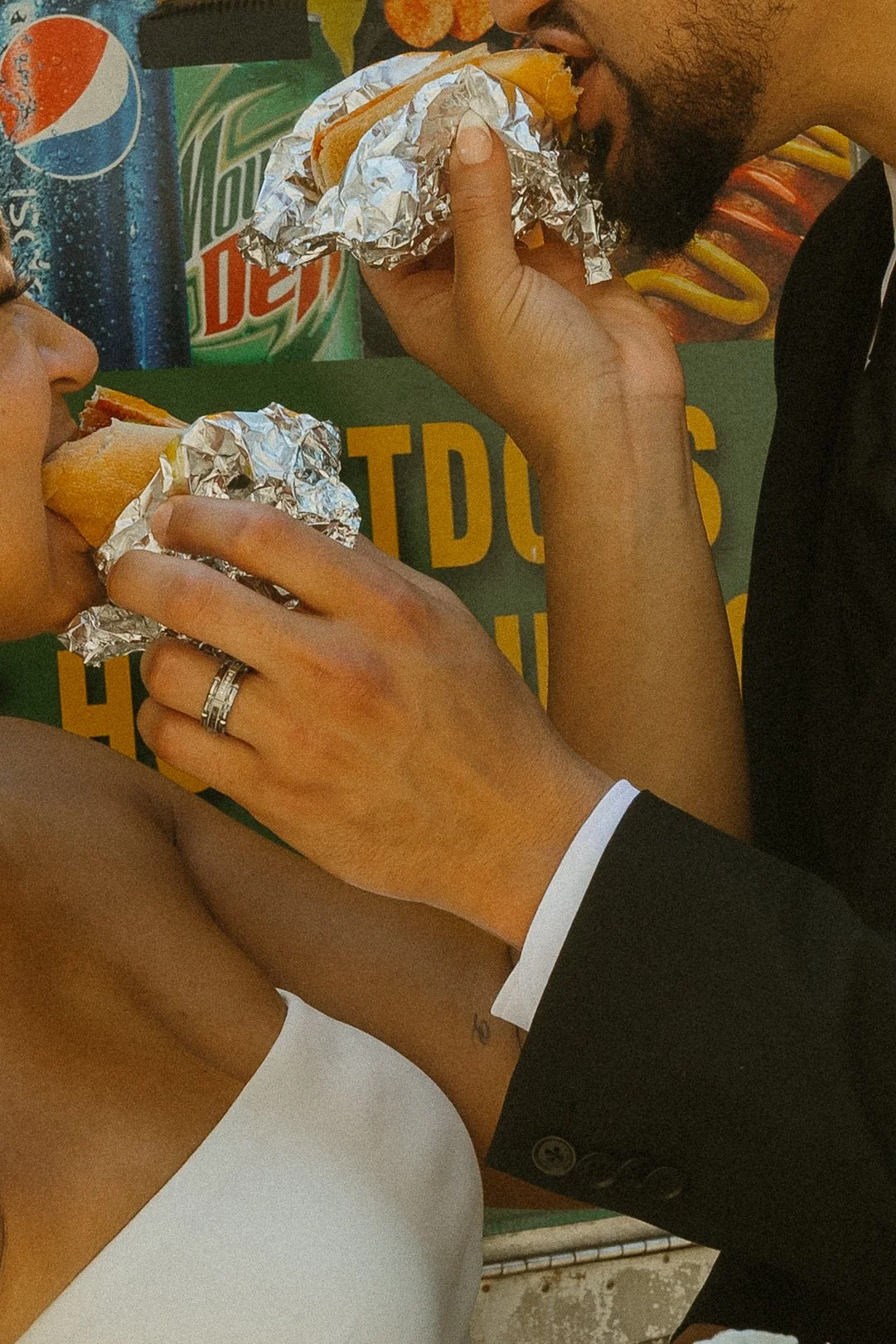A couple in formal attire eating hot dogs wrapped in aluminum foil at an outdoor event with soda cans and a colorful backdrop.