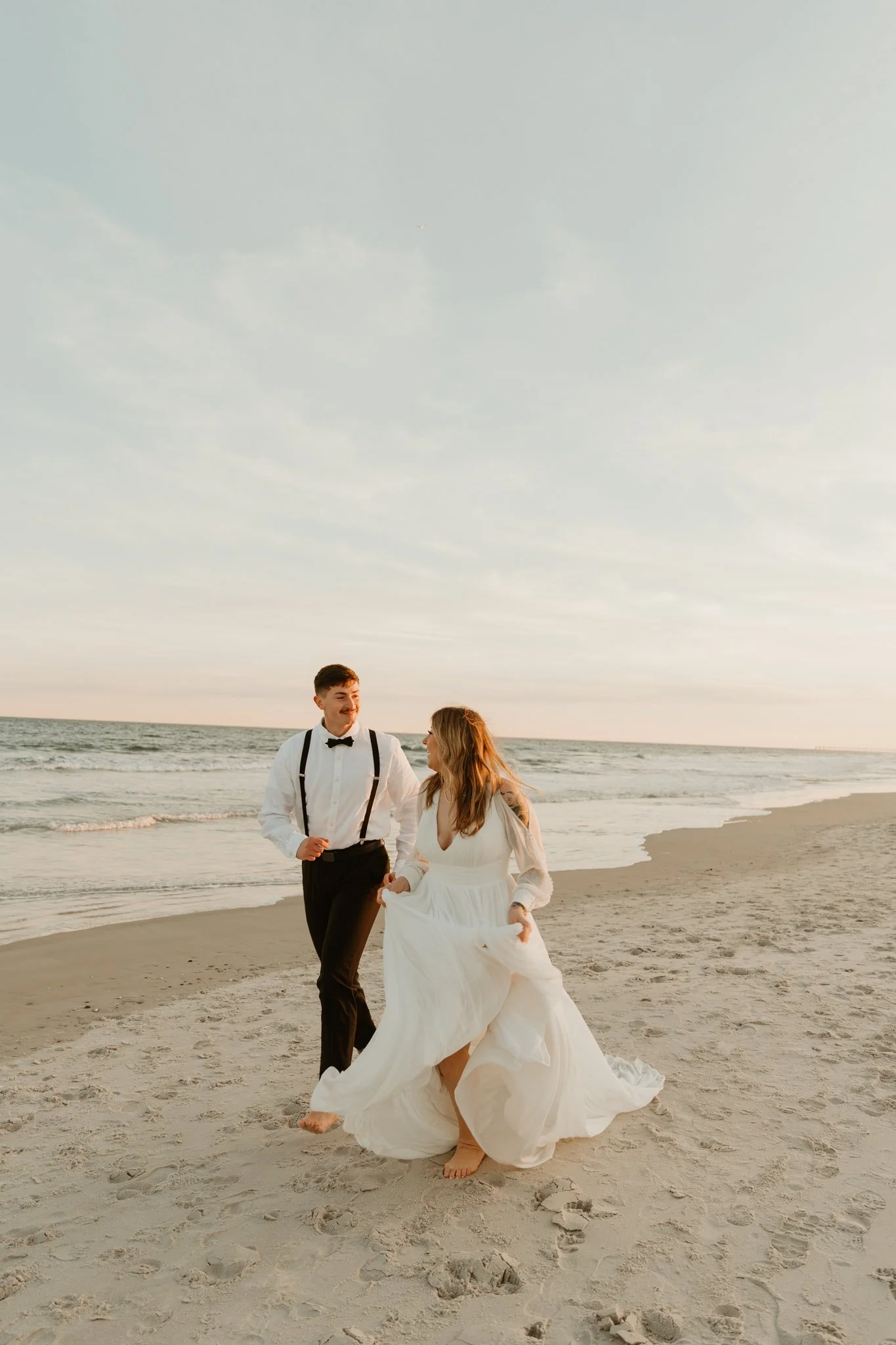 A couple dressed in wedding attire walking barefoot on a beach during sunset, with the ocean and sky in the background.
