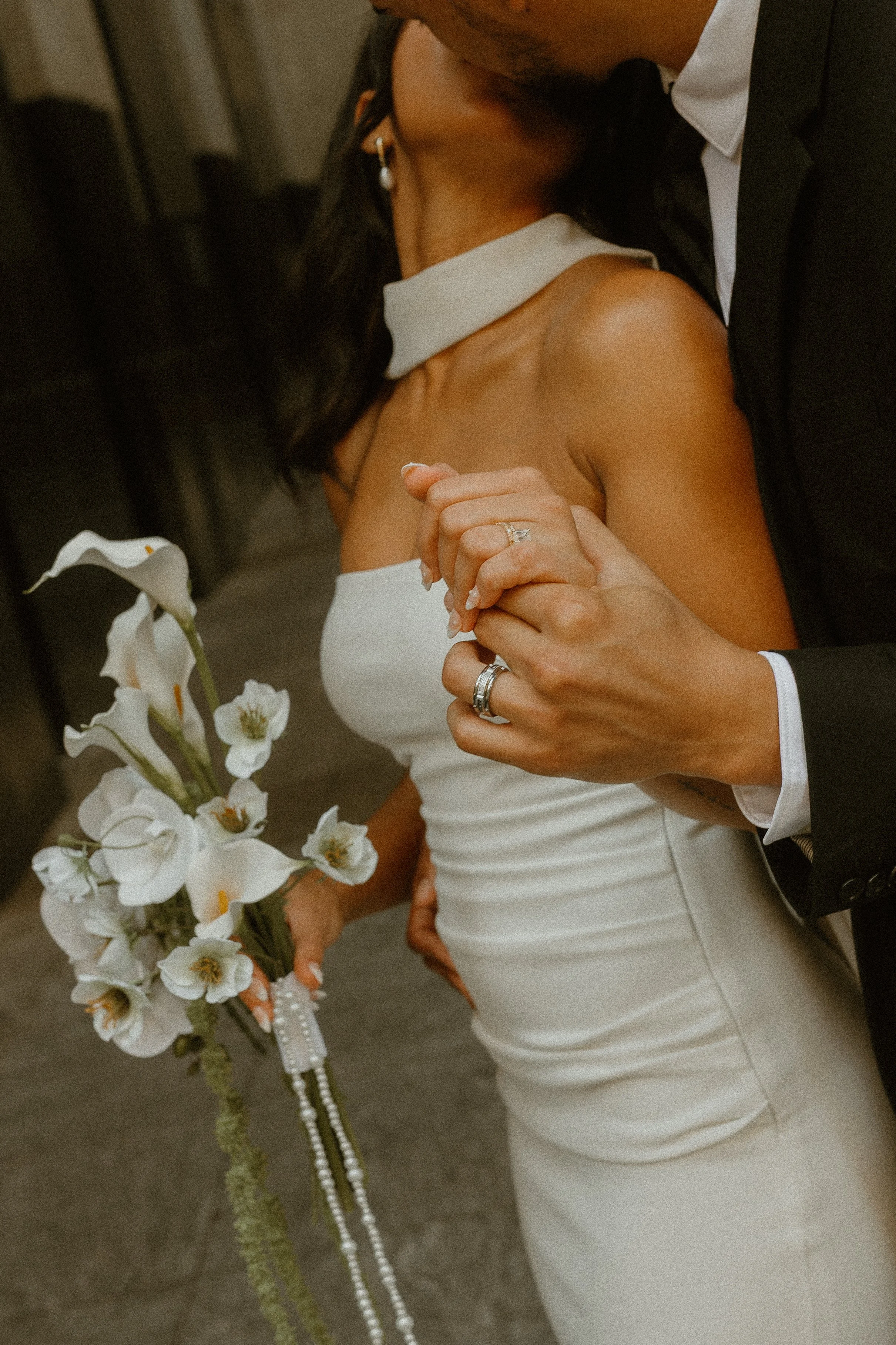 Close-up of a bride and groom in an intimate embrace, with the bride holding a white orchid bouquet and wearing a white strapless wedding dress. The groom is dressed in a dark tuxedo, and they are holding hands with wedding rings visible. The focus i