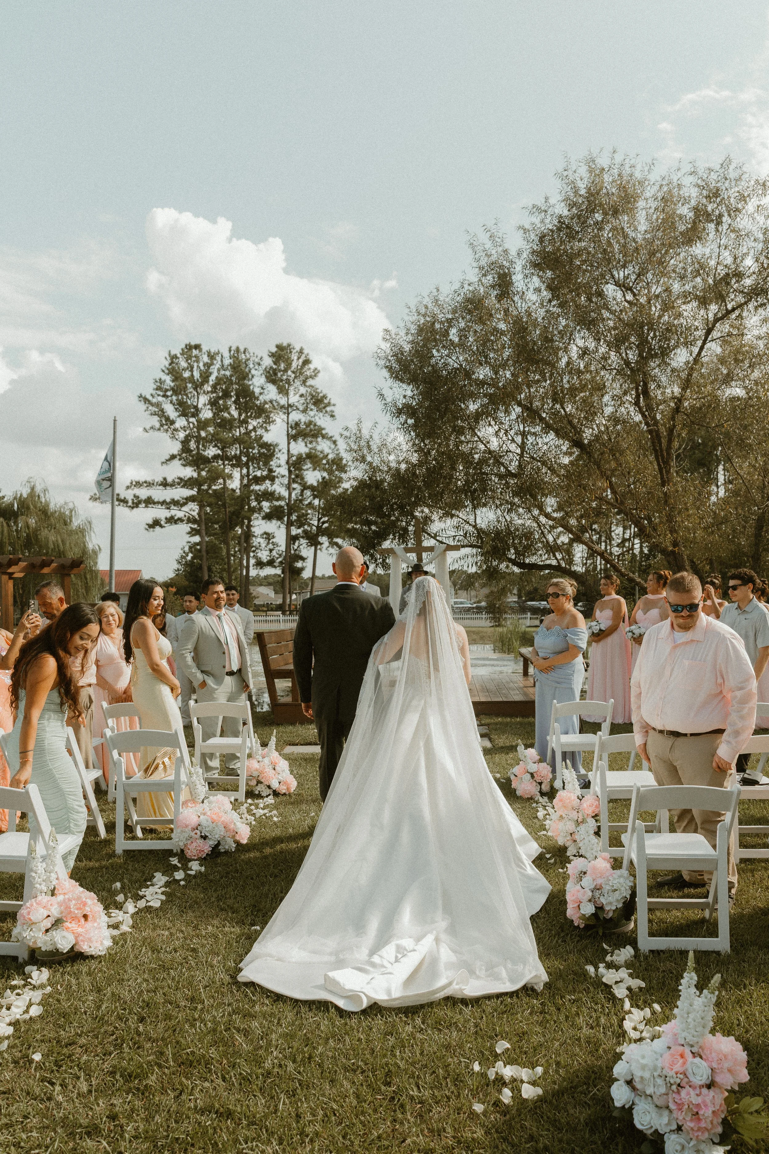 Outdoor wedding ceremony with a bride in a white gown and a groom in a black suit standing at an altar, surrounded by guests seated on white chairs decorated with pink and white flowers, under a large tree on a grassy lawn.