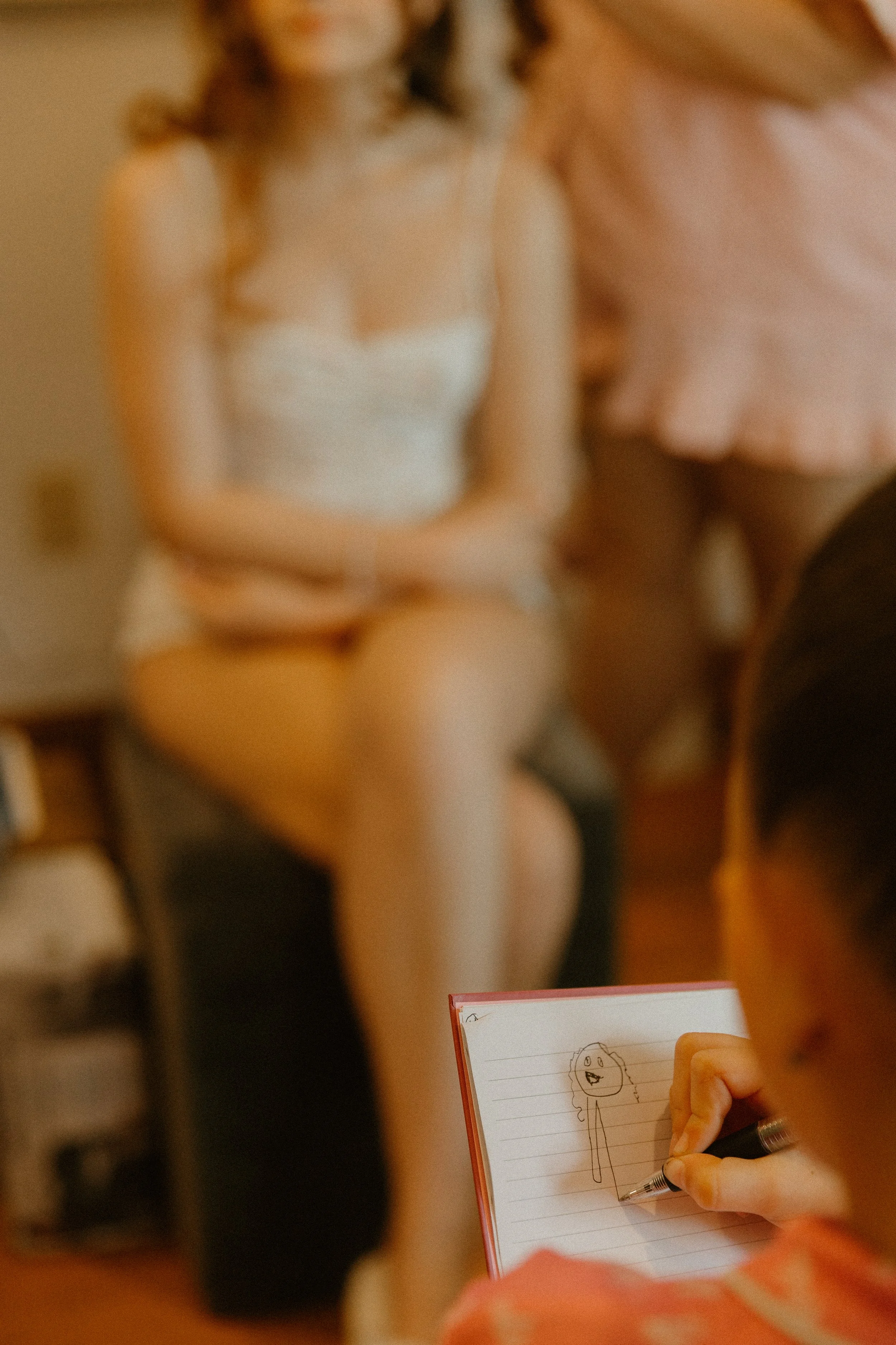 A person is drawing a stick figure with a round head and hair on a notepad, while another person is blurred in the background.