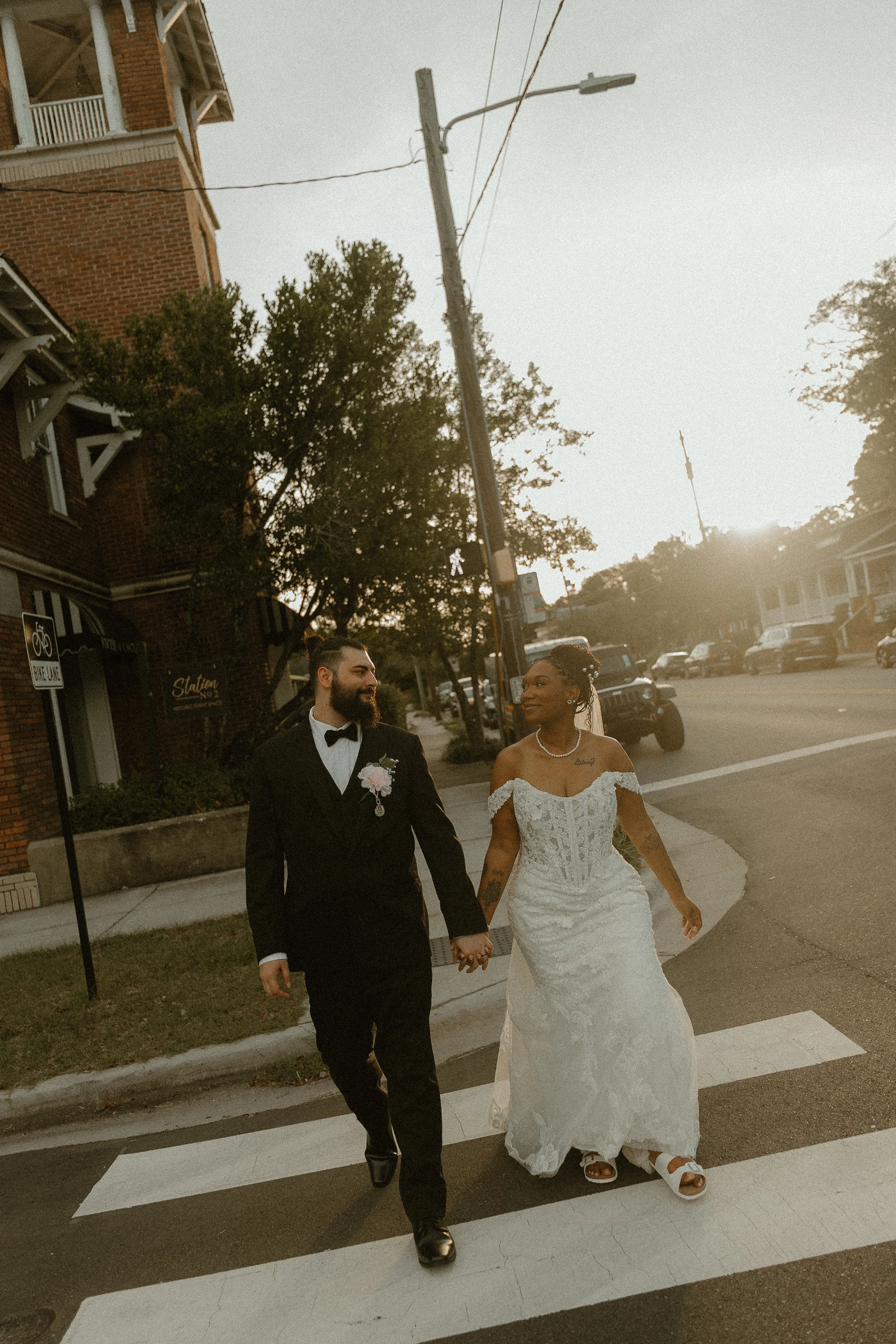 A newlywed couple, the groom dressed in a tuxedo and the bride in a white wedding dress, walk hand in hand across a crosswalk on a city street during sunset.