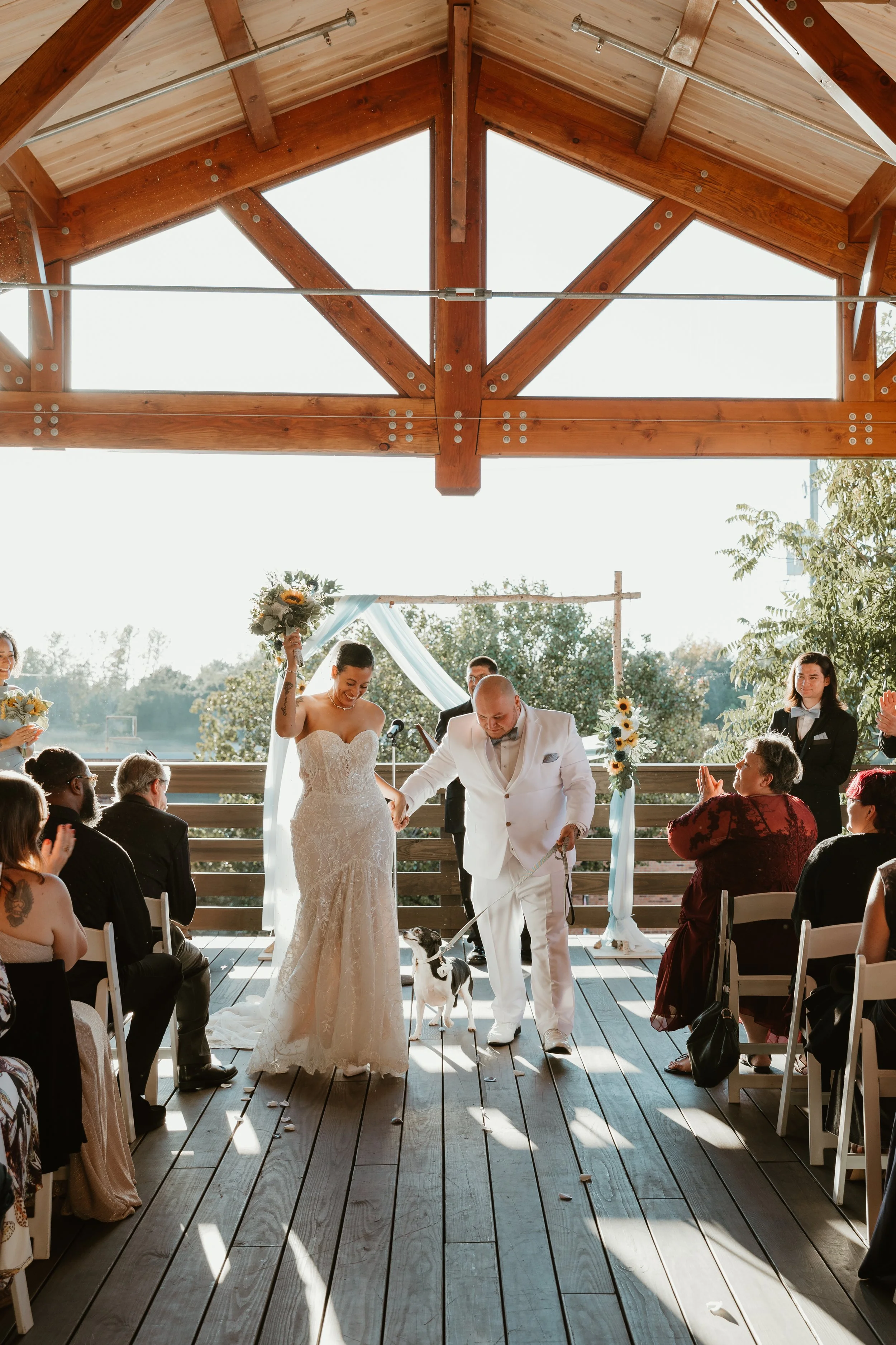 A wedding ceremony with a bride in a white lace gown holding a bouquet and a groom in a white suit with a small dog. Guests are seated on either side, clapping and smiling, under a wooden pavilion with sunlight streaming in.