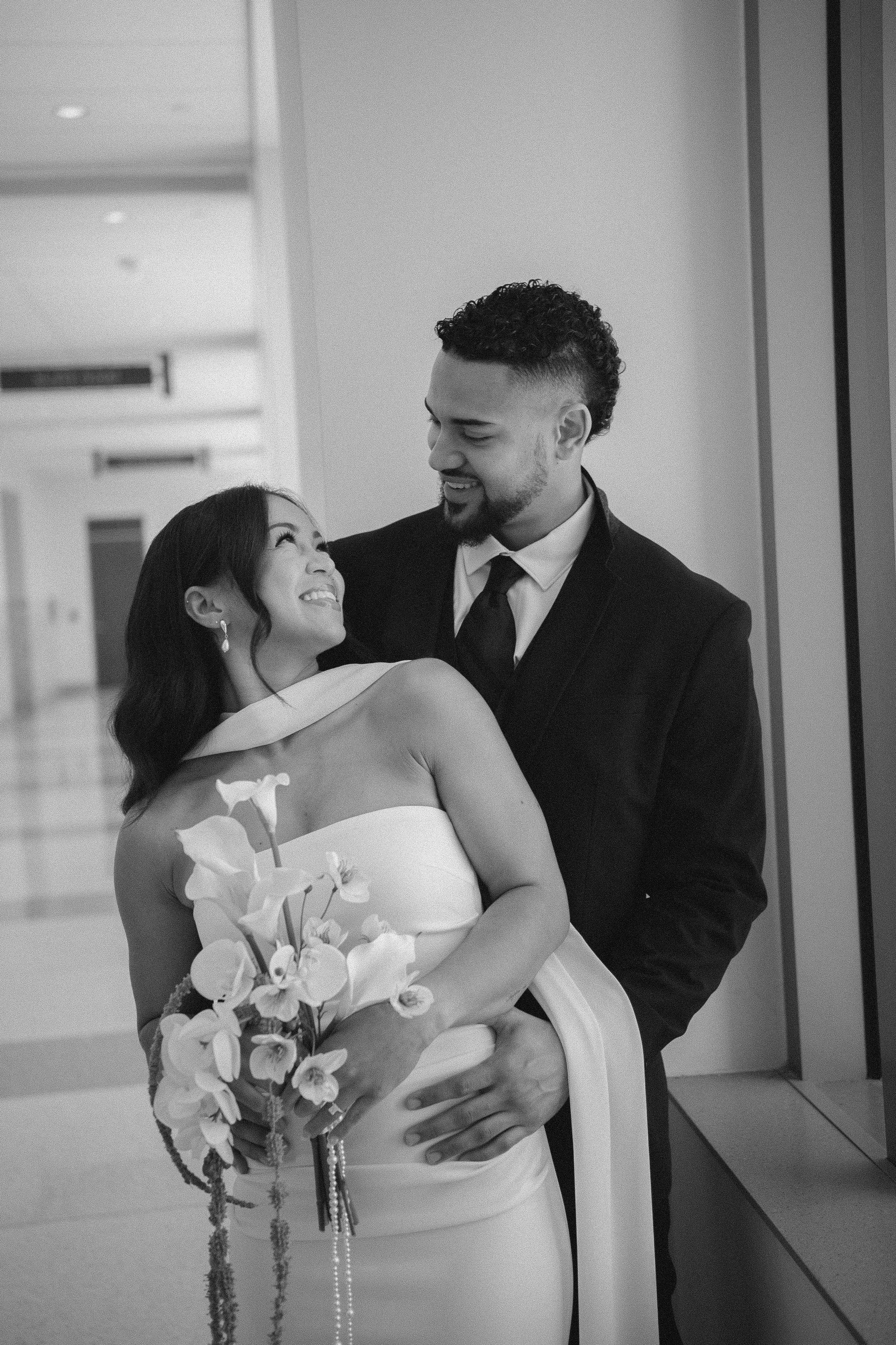 A couple dancing closely at a wedding reception. The woman is wearing a sleeveless wedding gown with floral embellishments, holding a bouquet of flowers, and has long dark hair. The man is wearing a dark suit with a tie, has short curly hair, and is smiling as he looks at her.