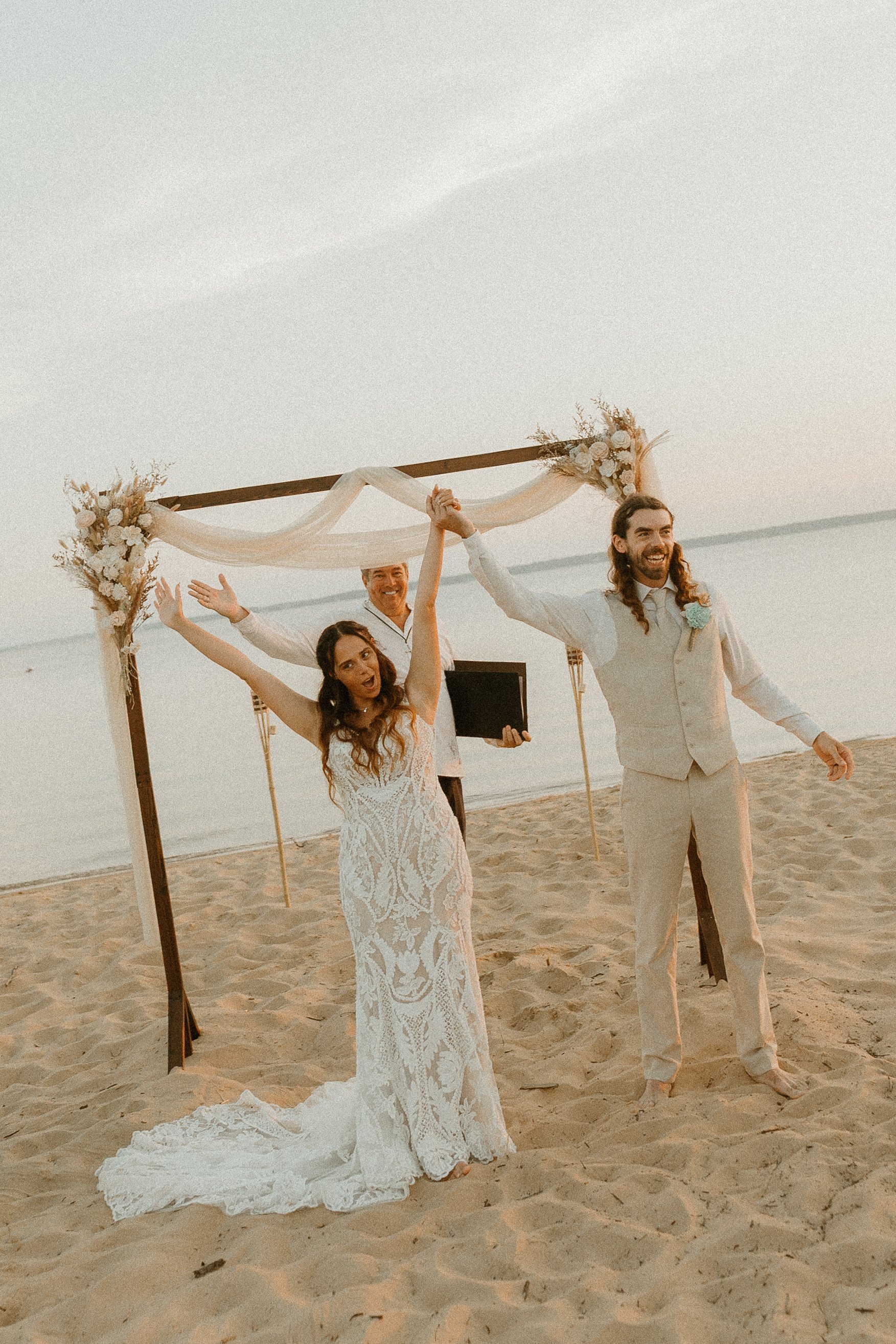 A wedding ceremony on the beach with a couple, the bride in a white lace dress and the groom in light-colored pants and a vest, smiling and holding hands under a decorated wooden arch with flowers and fabric.