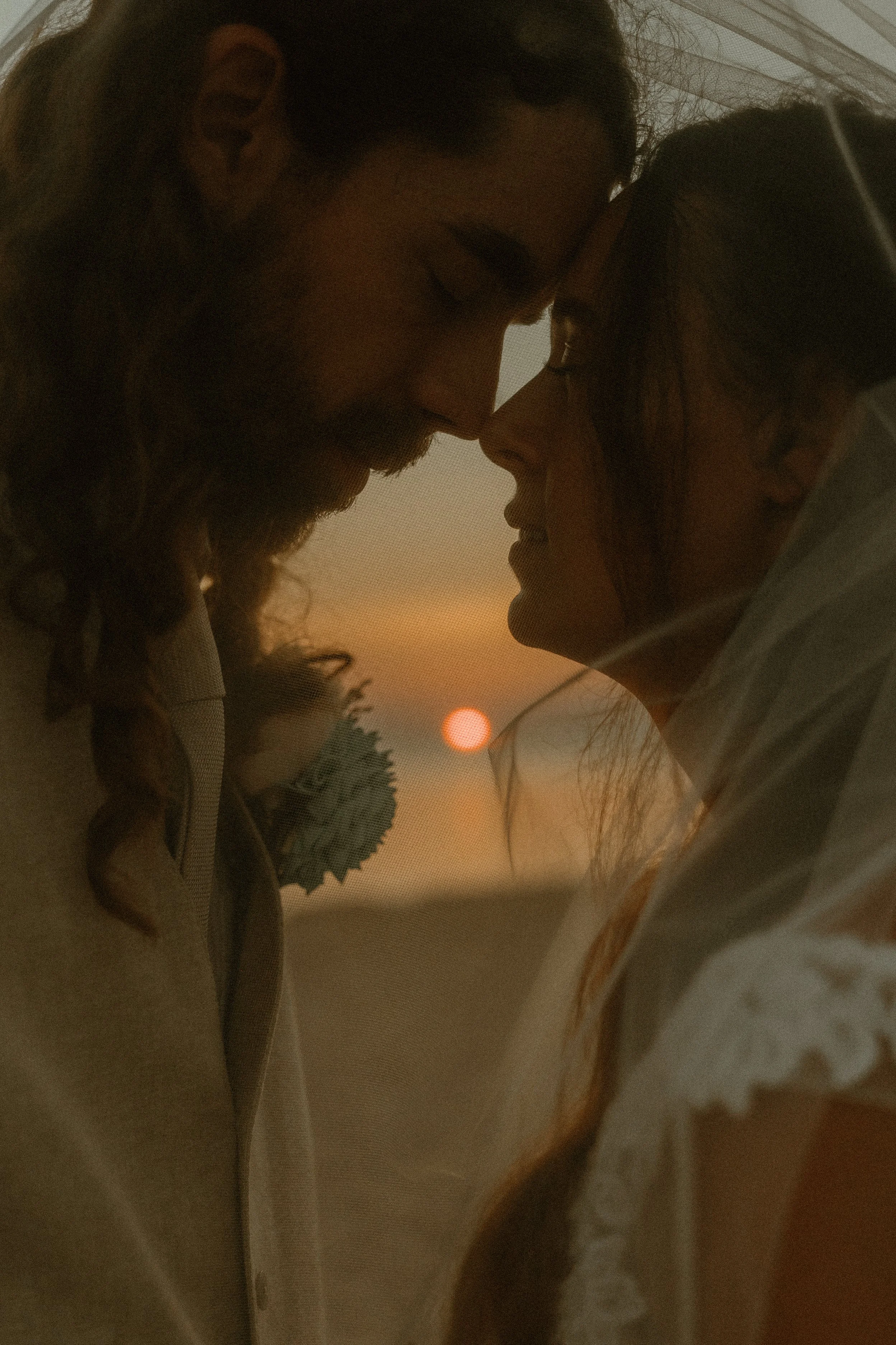 Close-up of a man and woman with foreheads touching, eyes closed, at sunset.