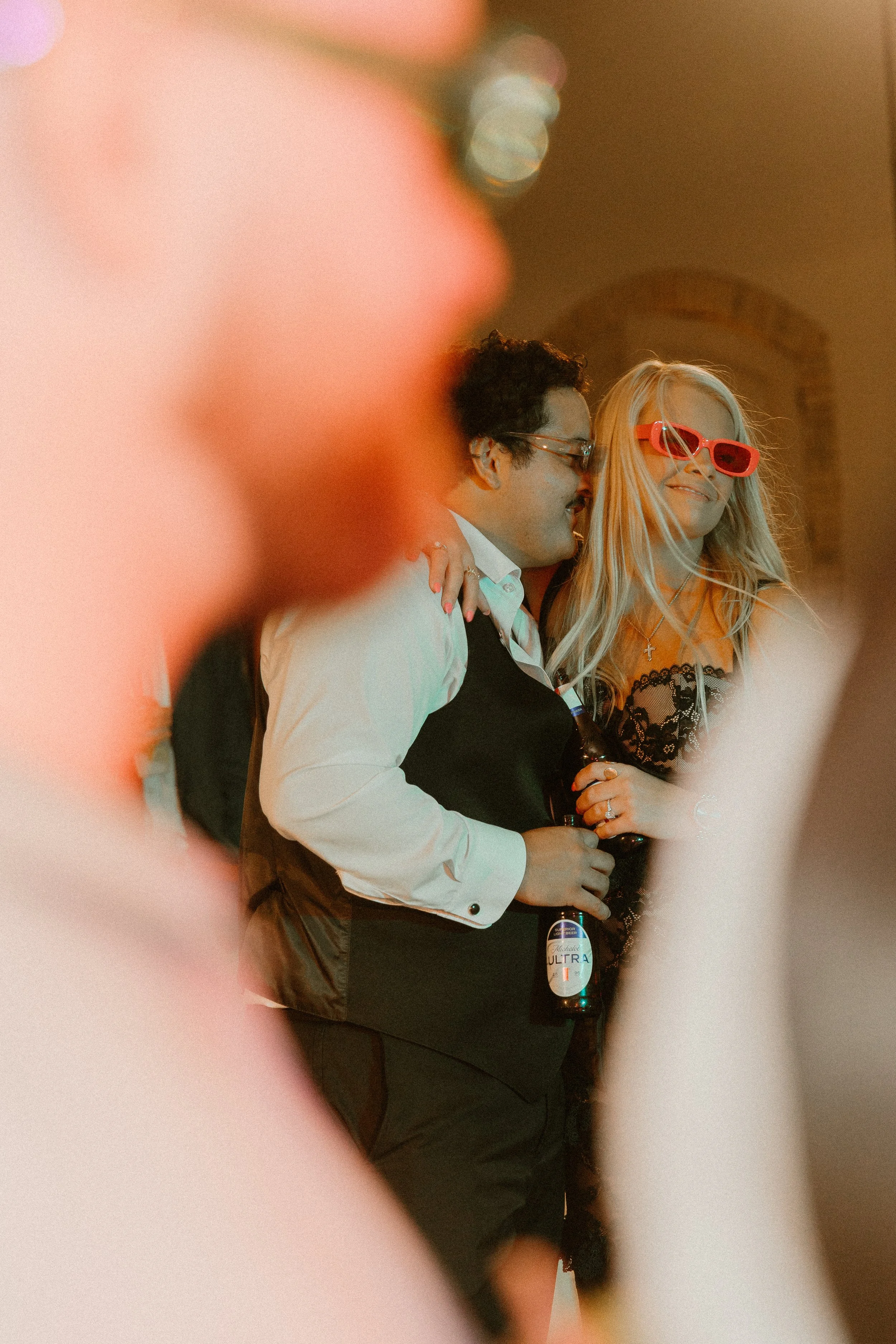 People at a social gathering, two women and a man smiling and holding drinks, one woman wearing red sunglasses, partially obscured by blurred foreground.