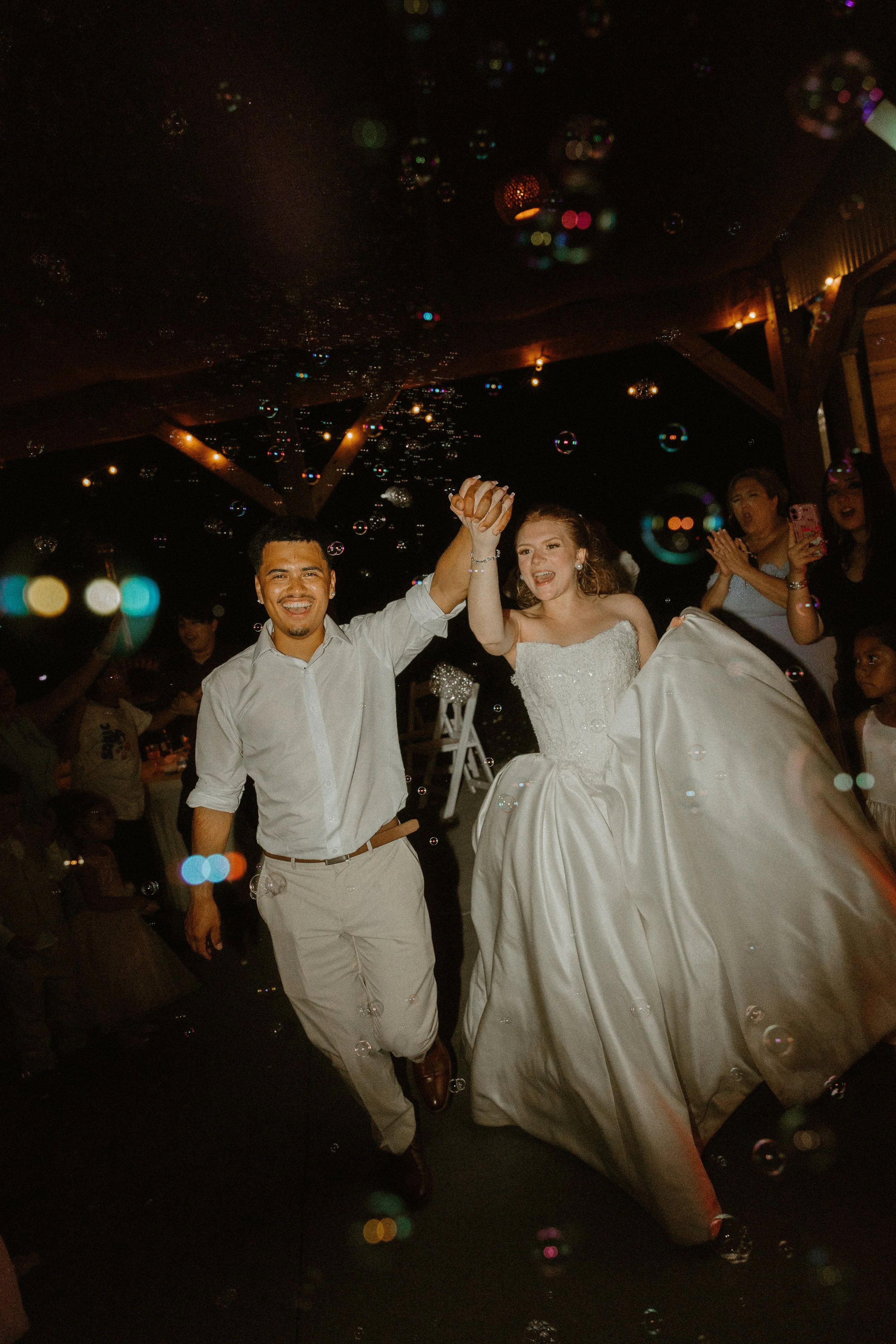 A newlywed couple holding hands and smiling at their wedding reception at night, with friends and family around them, and bubbles floating in the air.