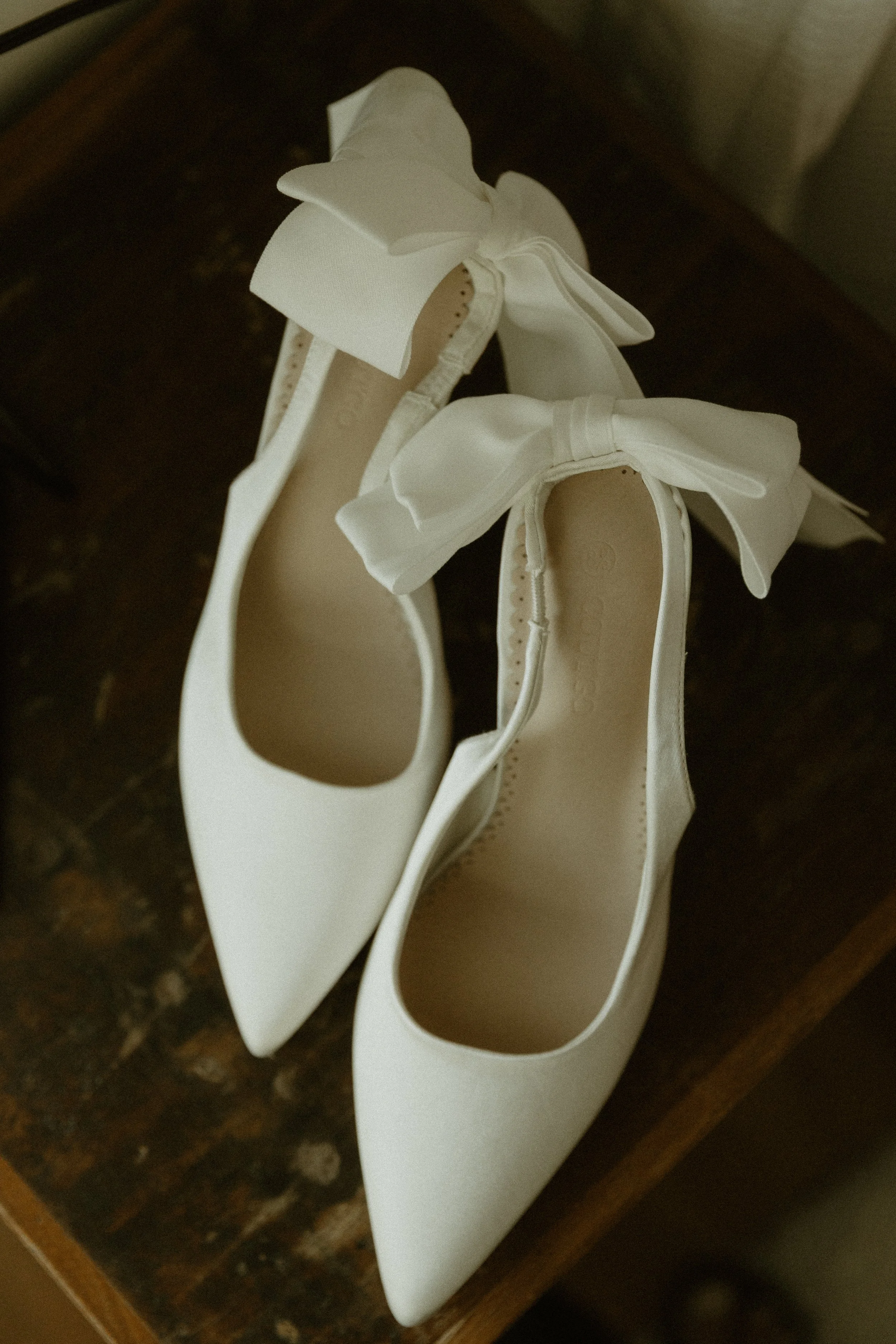 A pair of white women's ballet flats with large bows on the toes, placed on a dark wooden surface.