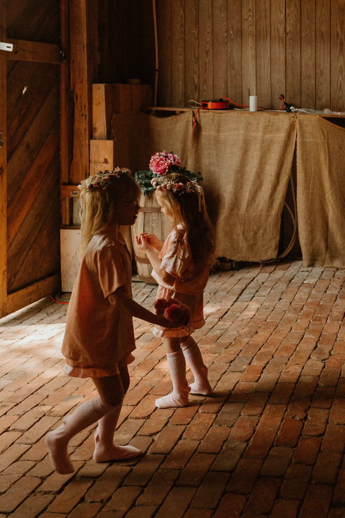 Two young girls wearing flower crowns and matching peach-colored outfits dance and smile in a rustic wooden barn with brick flooring.