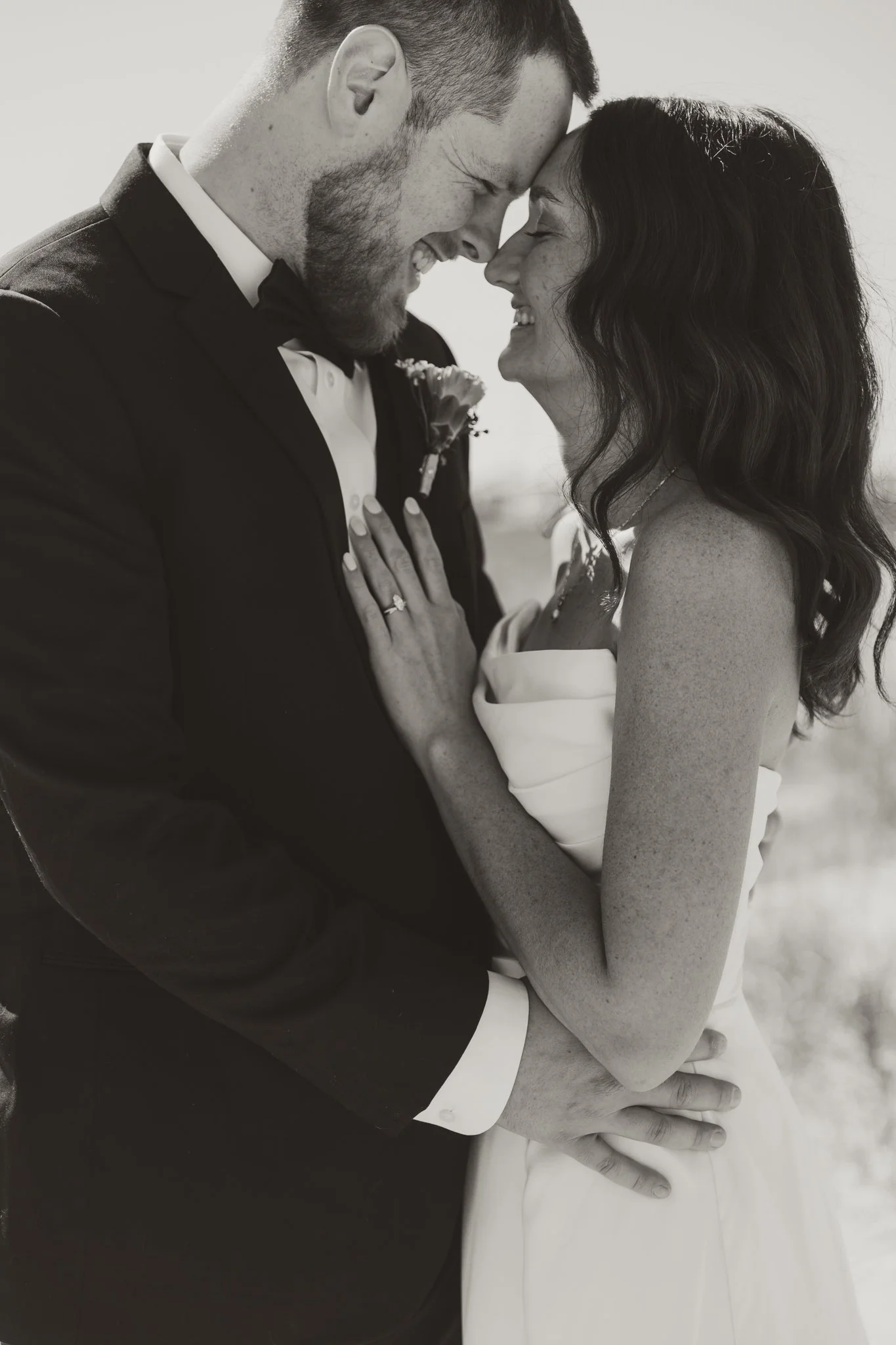 A black and white photo of a newlywed couple smiling and leaning their foreheads together, with the man in a tuxedo and the woman in a strapless wedding dress, sharing an intimate moment outdoors.