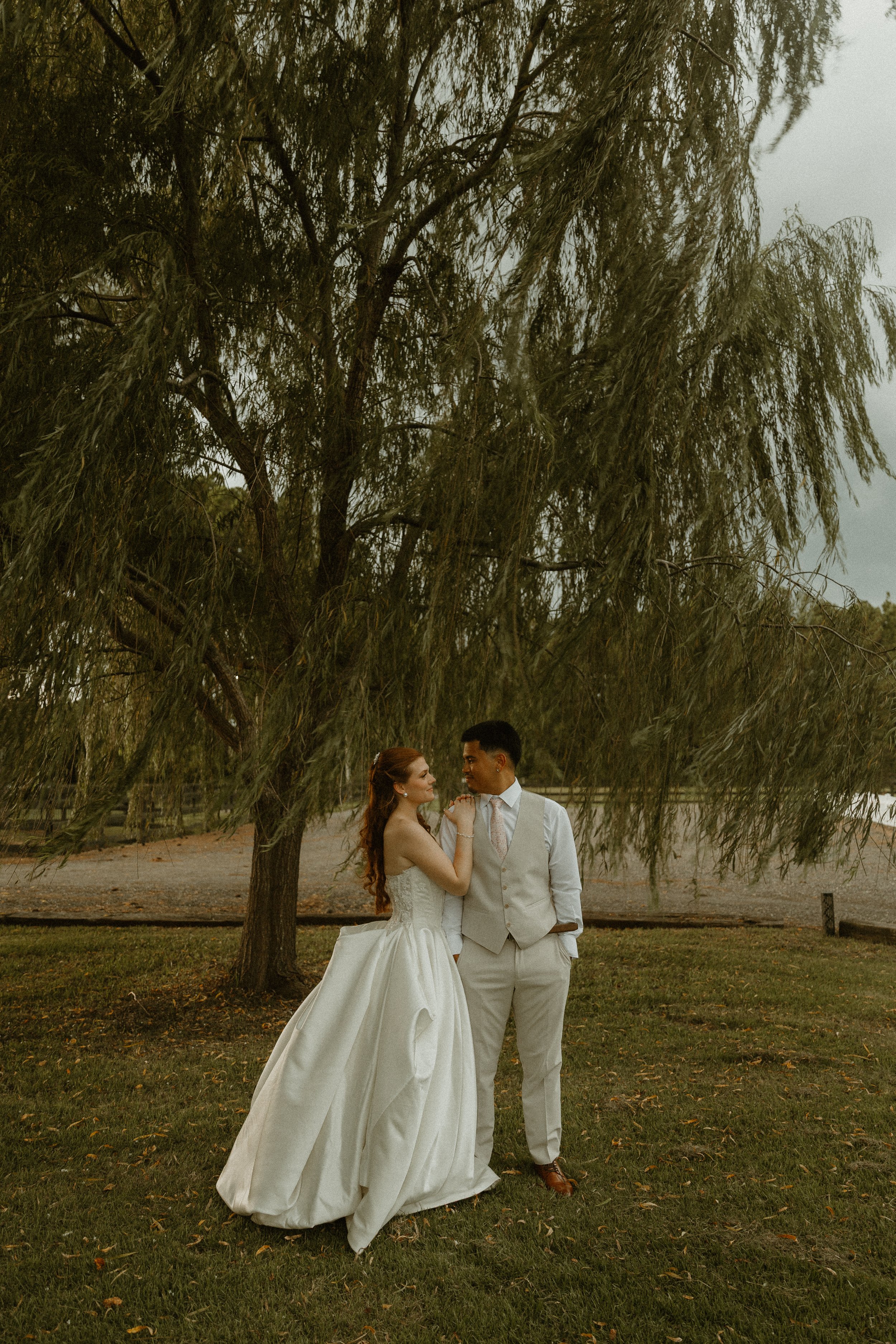 A bride and groom standing under a large tree in a park during daytime, with the bride wearing a white wedding dress and the groom in a beige vest and pants, holding hands and gazing at each other.